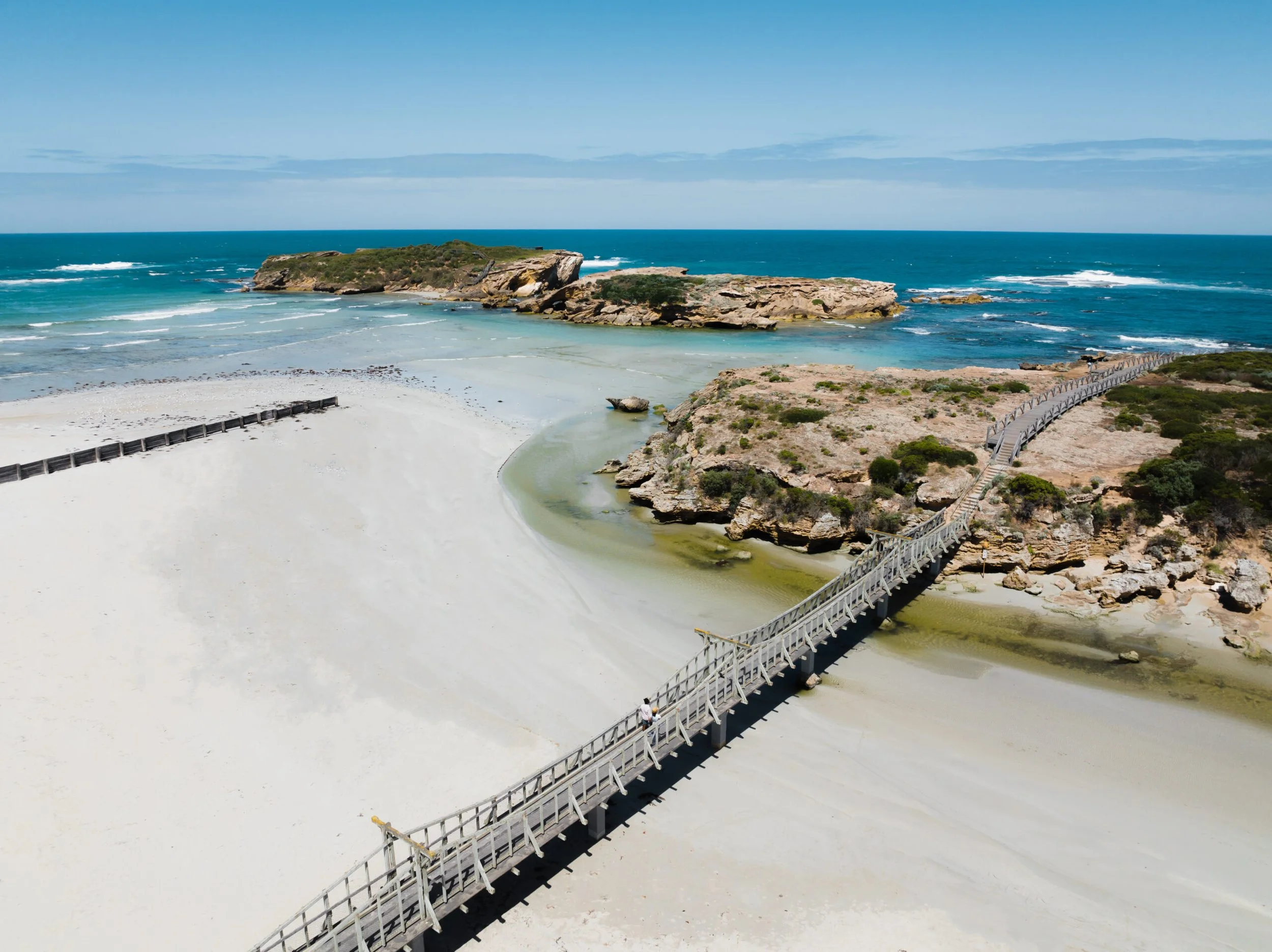 A wooden boardwalk crossing sandy beach and rocky cliffs overlooking the ocean under a blue sky.
