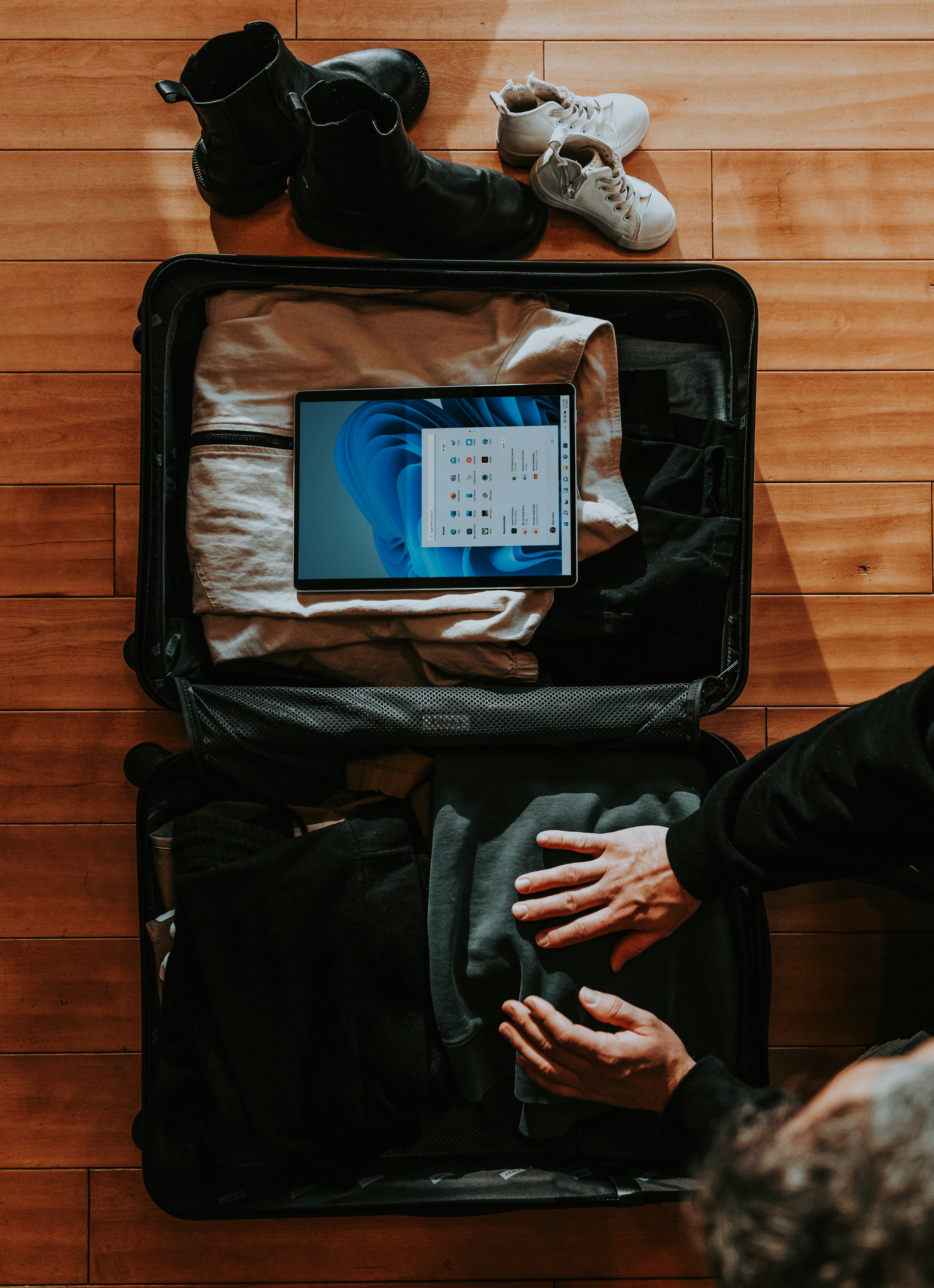 An open suitcase on a wooden floor filled with clothing, a tablet, and a person's hand packing clothes into it. Nearby are black boots and white sneakers.