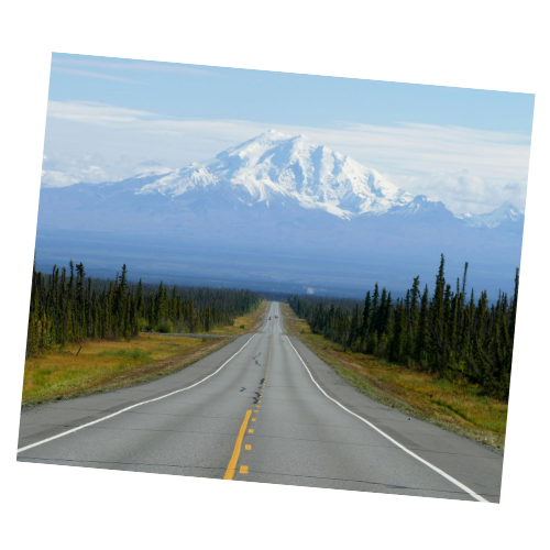A straight road stretches towards a snow-capped mountain, flanked by green trees on both sides under a partly cloudy sky.