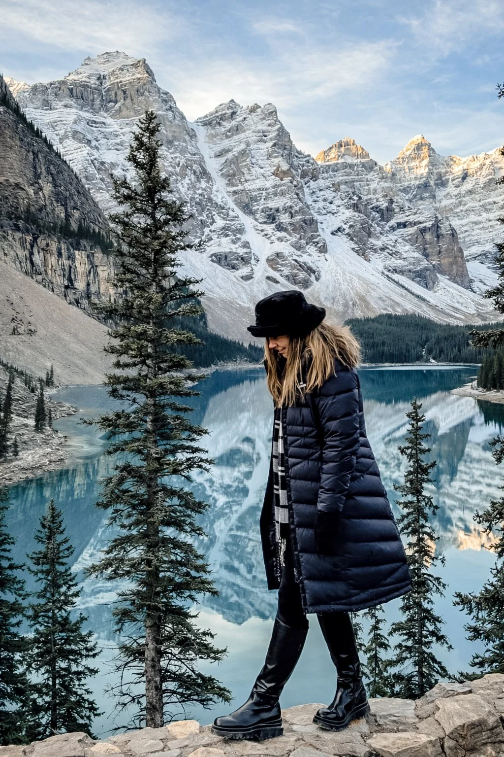 A woman standing on rocks near a lake with snow-capped mountains and evergreen trees in the background.