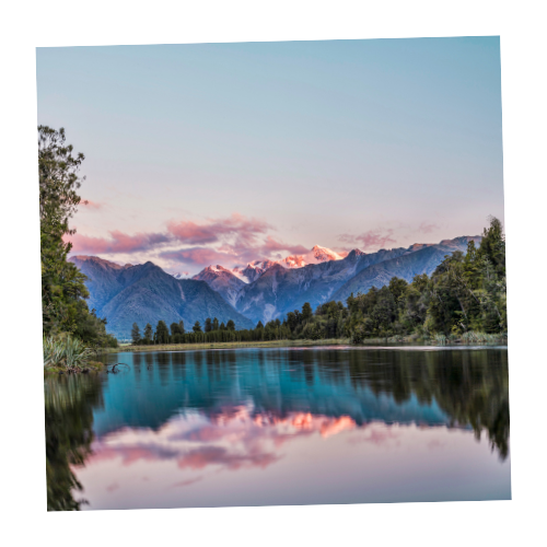 Serene lake surrounded by trees with mountain range in the background under a pinkish sunset sky.