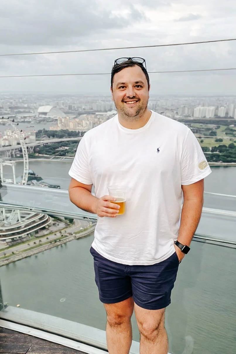 A man standing on a high observation deck holding a small cup of beer. He is wearing a white t-shirt with a small logo, dark shorts, and a watch. The background features a city view with a river, a Ferris wheel, and modern buildings under a cloudy sky.