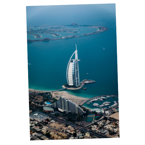 An aerial view of the Burj Al Arab hotel shaped like a sail, located on an artificial island off the coast of Dubai, with surrounding water and cityscape.
