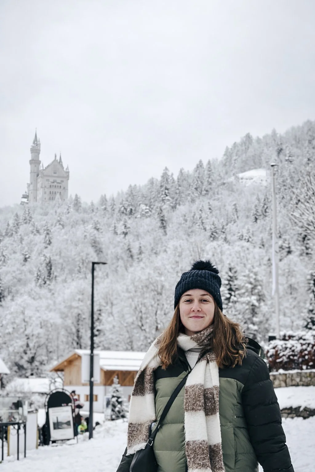 A woman dressed in winter clothing, including a black beanie and a striped scarf, standing outdoors in a snowy landscape with snow-covered trees, a chalet-style building, and a castle on a hill in the background.