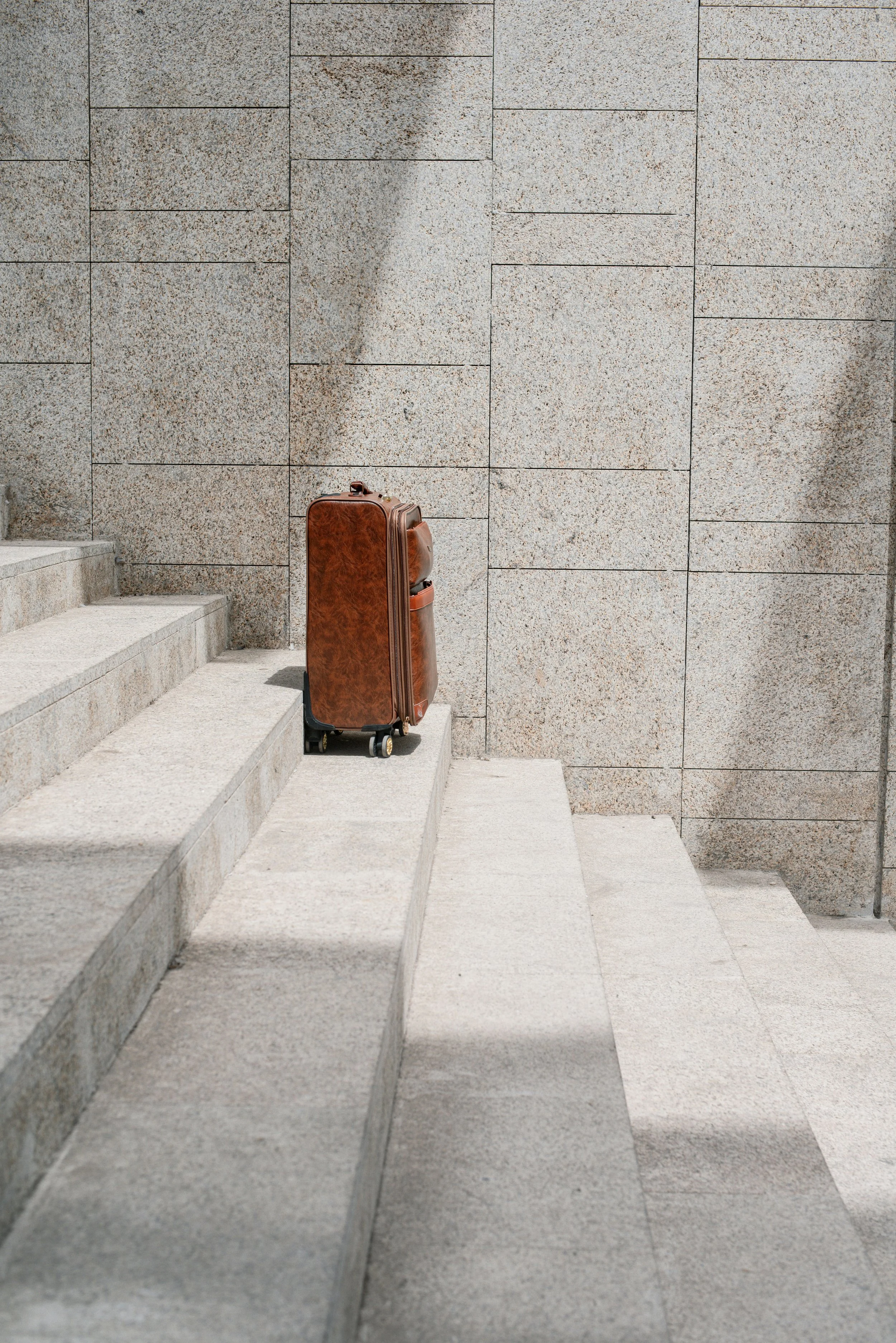 A brown rolling suitcase resting on a stone staircase against a textured stone wall.