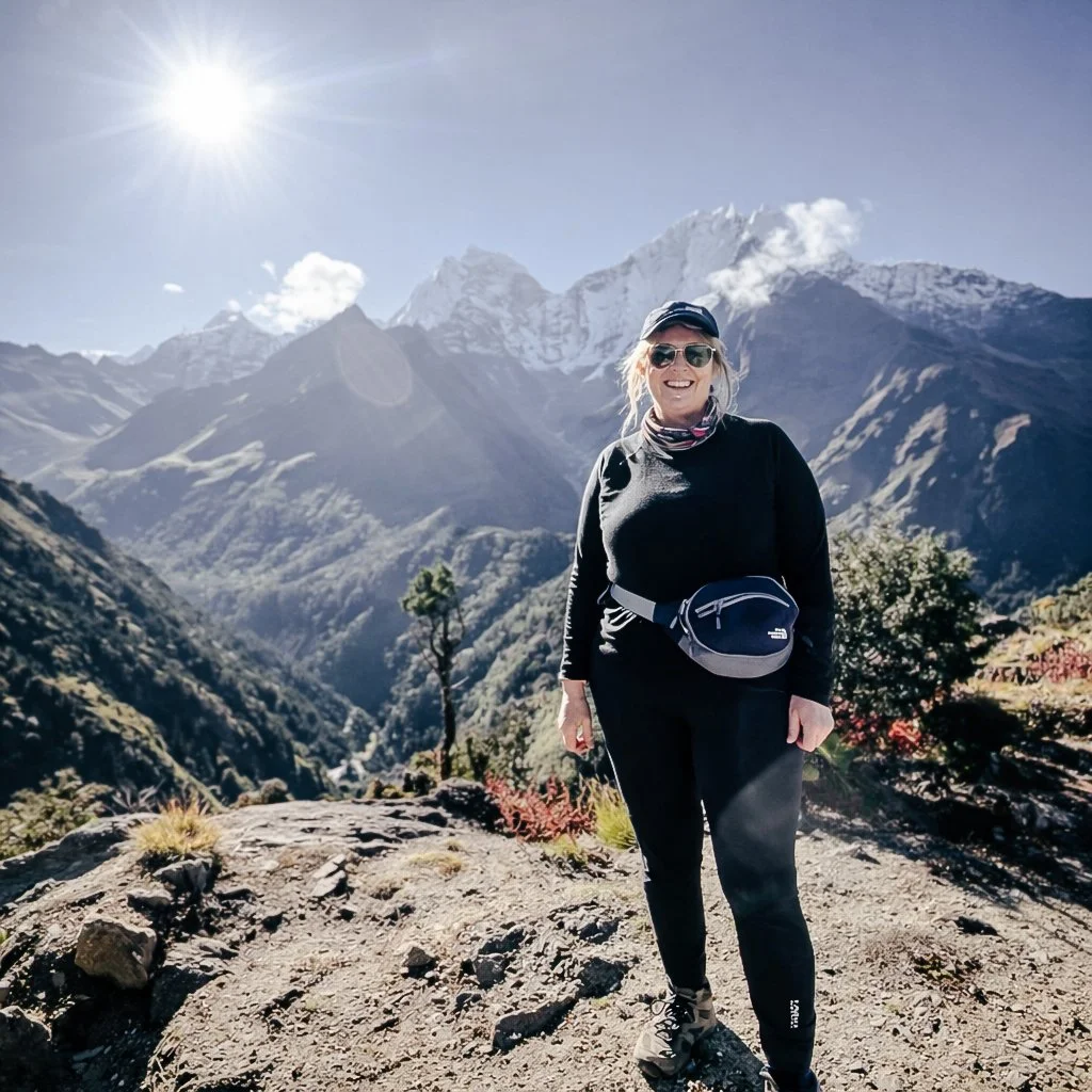 A woman wearing sunglasses, a black long-sleeve shirt, and hiking gear stands on a rocky trail in a mountainous landscape with snow-capped peaks and a bright sun overhead.
