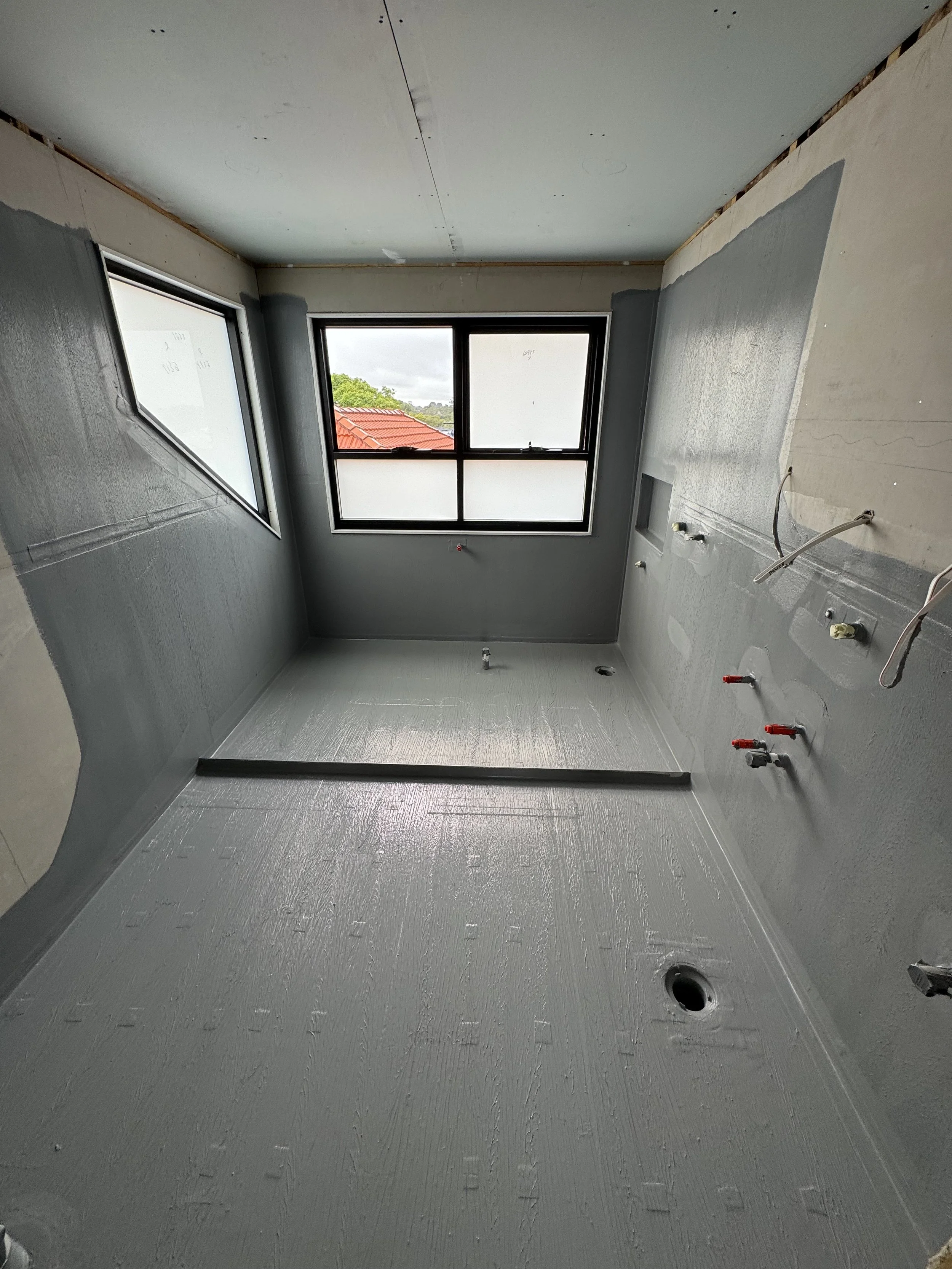 Empty bathroom under construction with gray waterproofing on walls and floor, plumbing fixtures not yet installed, and large windows with views outside.