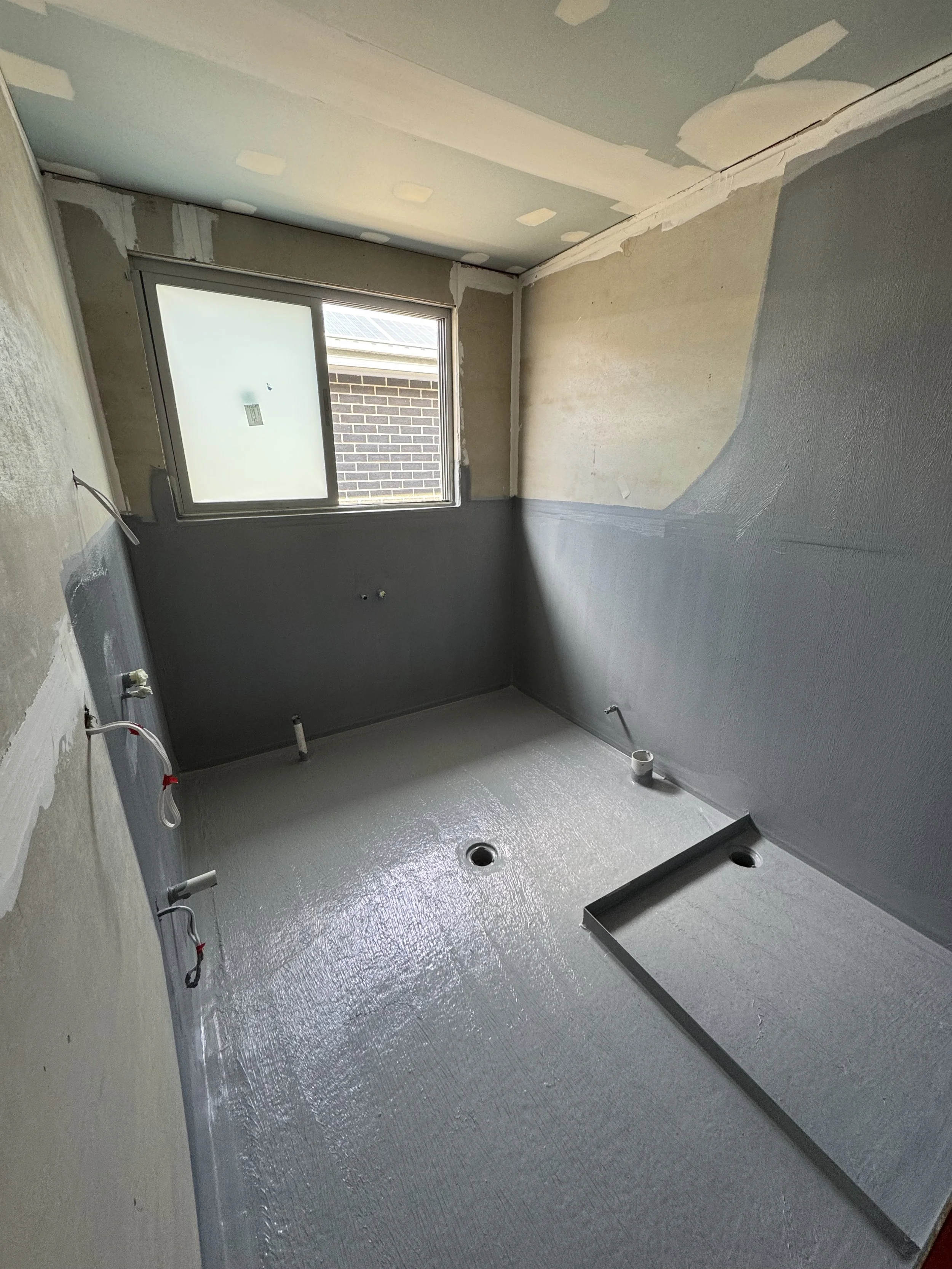 Room under construction with gray waterproofing on the floor and lower walls, unfinished walls with drywall, and a window showing a brick building outside.