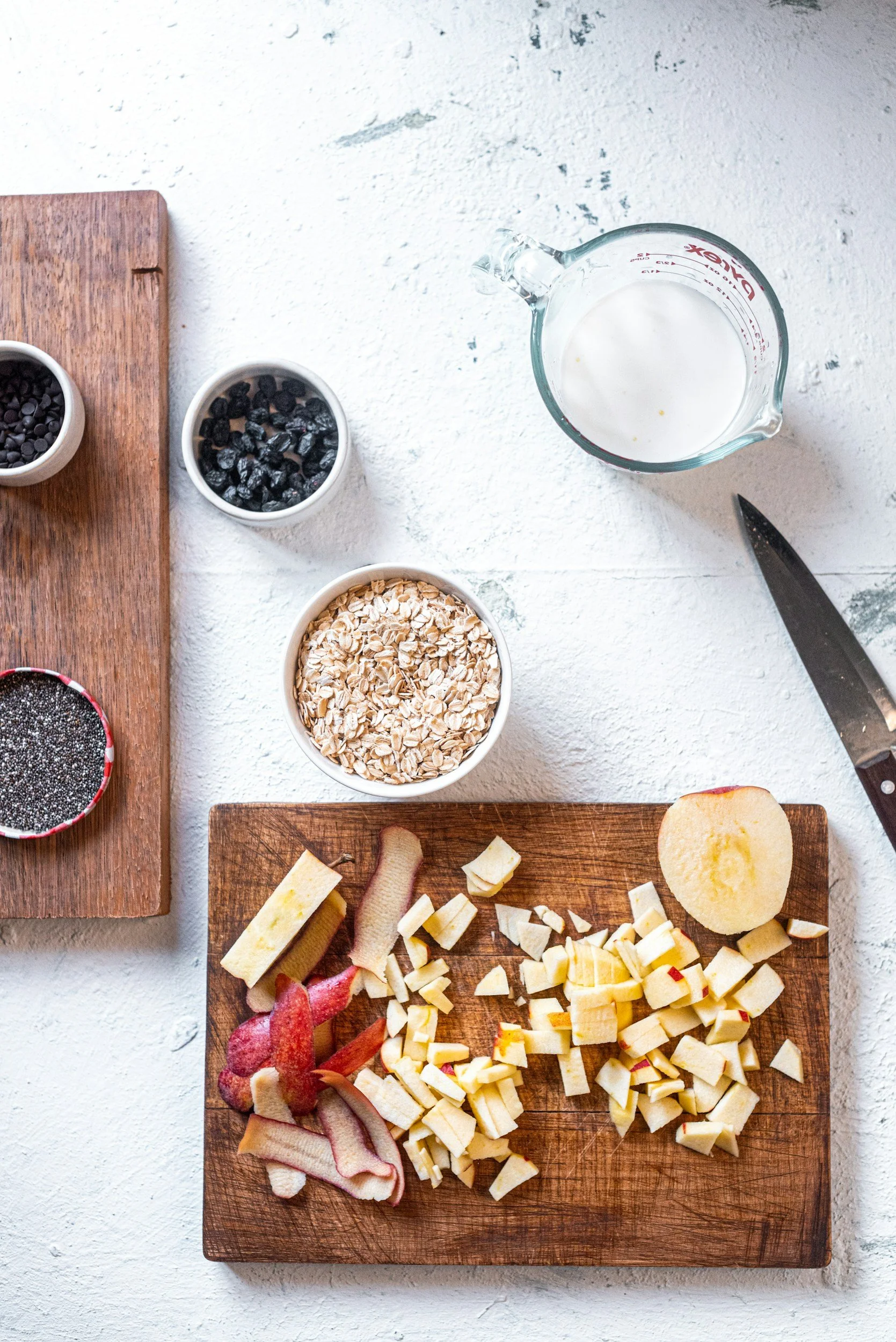 Wooden cutting board with peeled and chopped apples, a small bowl of oats, black and white seeds, milk in a glass measuring cup, and a kitchen knife on a white textured surface.