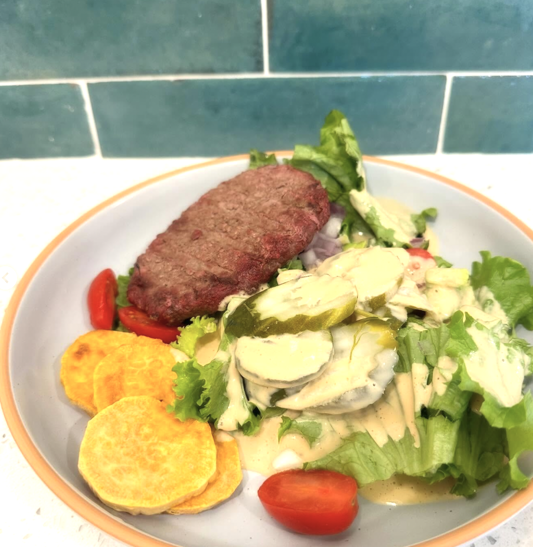 Plate with hamburger patty, salad with lettuce, cherry tomatoes, sliced cucumbers, and potato chips against a blue subway tile background.