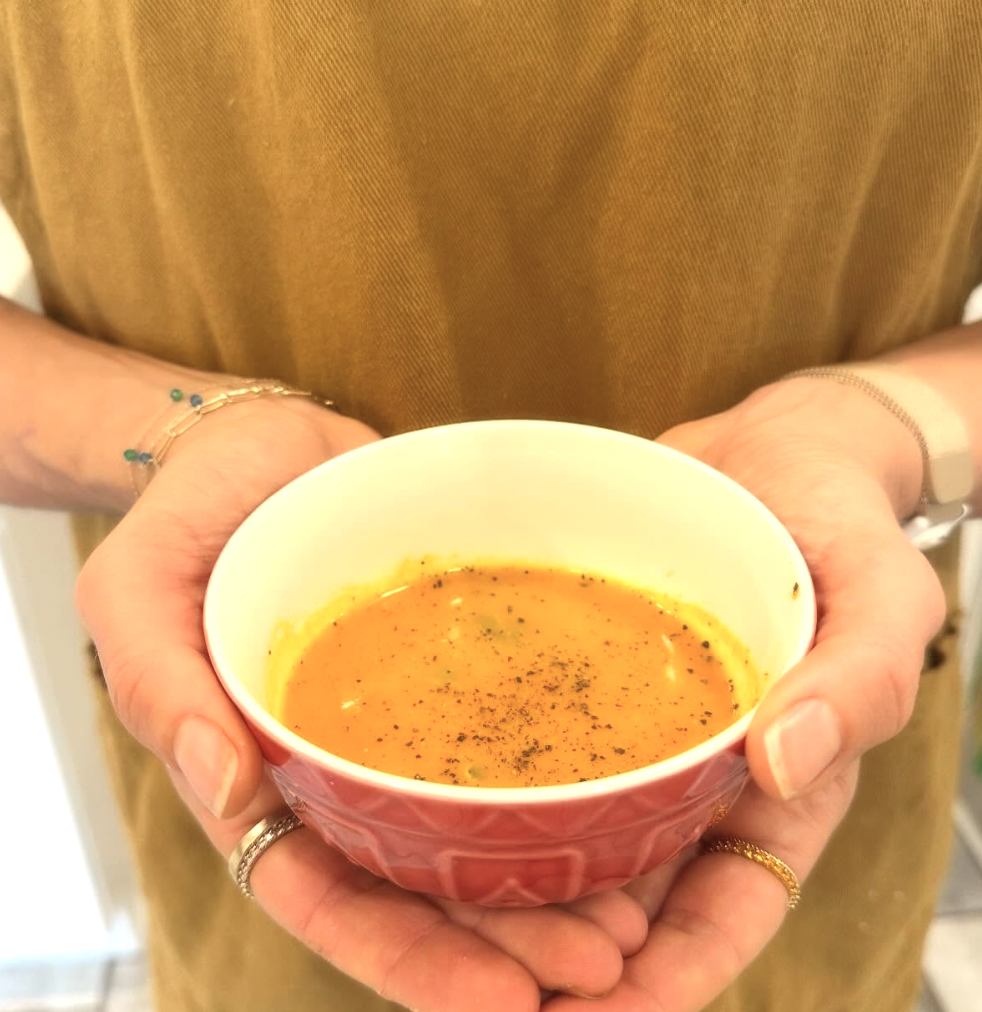 Person holding a small bowl of orange-colored soup garnished with herbs, wearing rings and bracelets.