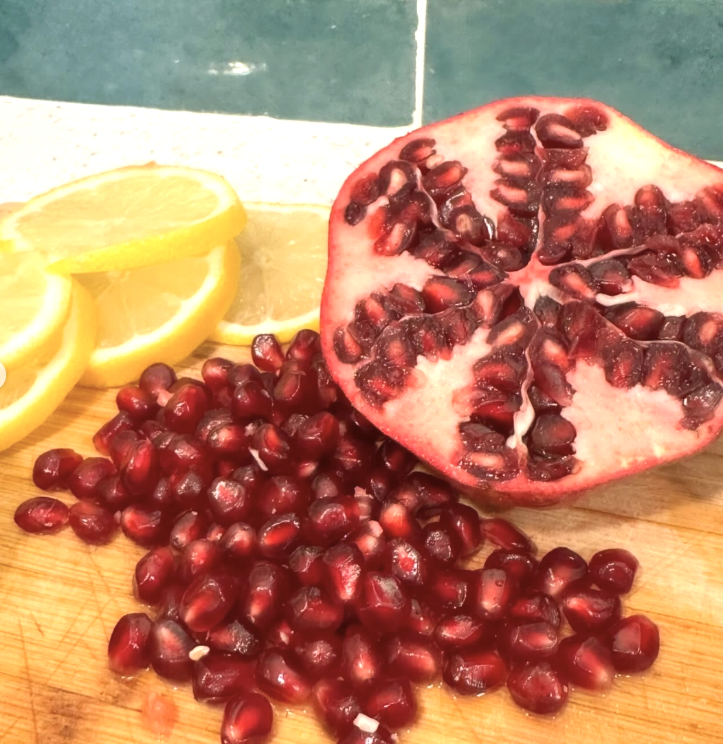 A halved pomegranate with seeds spilled on a wooden cutting board, and lemon slices in the background.