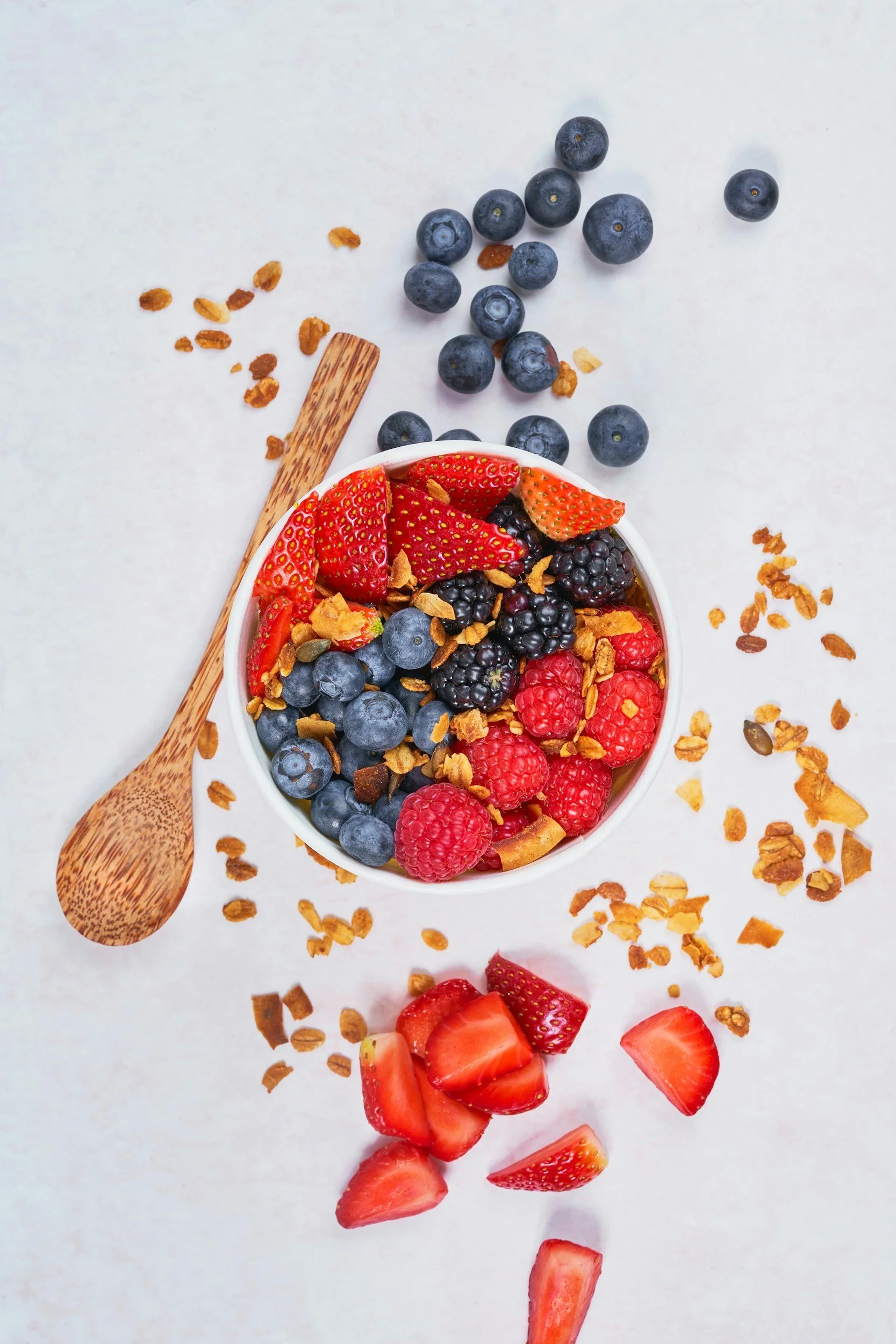 A white bowl filled with mixed berries, including strawberries, blueberries, blackberries, and raspberries, topped with granola. A wooden spoon rests beside the bowl, with scattered granola and loose berries on a white surface.