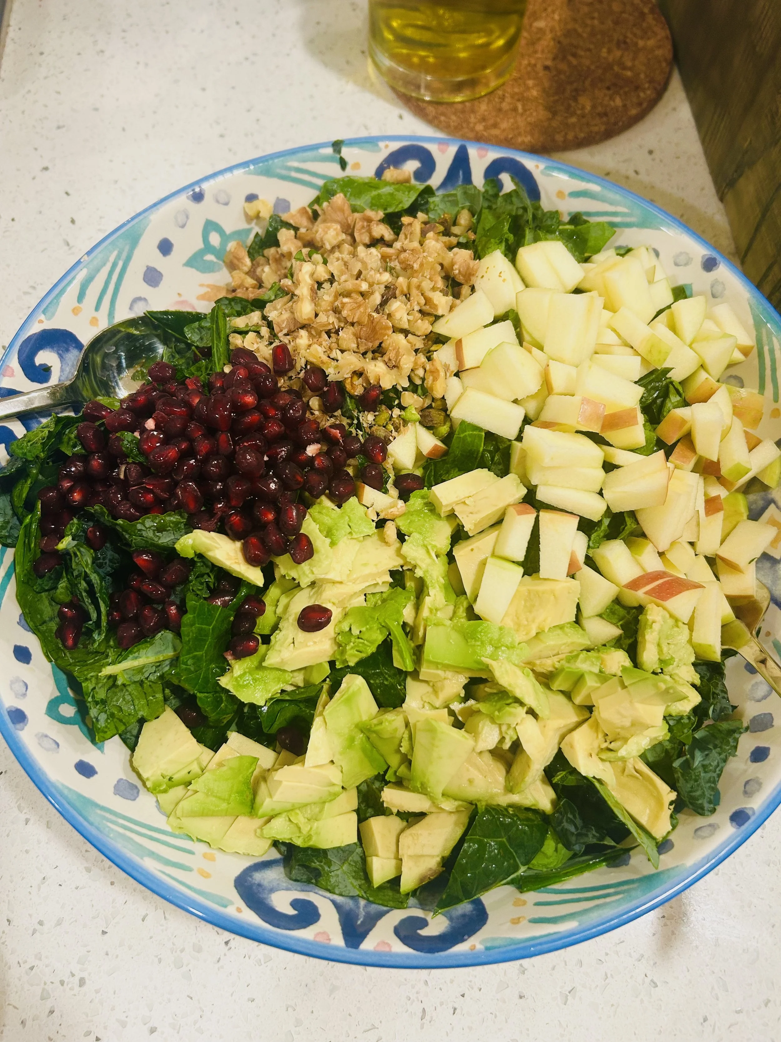 A colorful salad in a decorative bowl with chopped apples, avocado, walnuts, and pomegranate seeds. A spoon is resting in the salad, and a bottle of olive oil and a coaster are visible in the background.