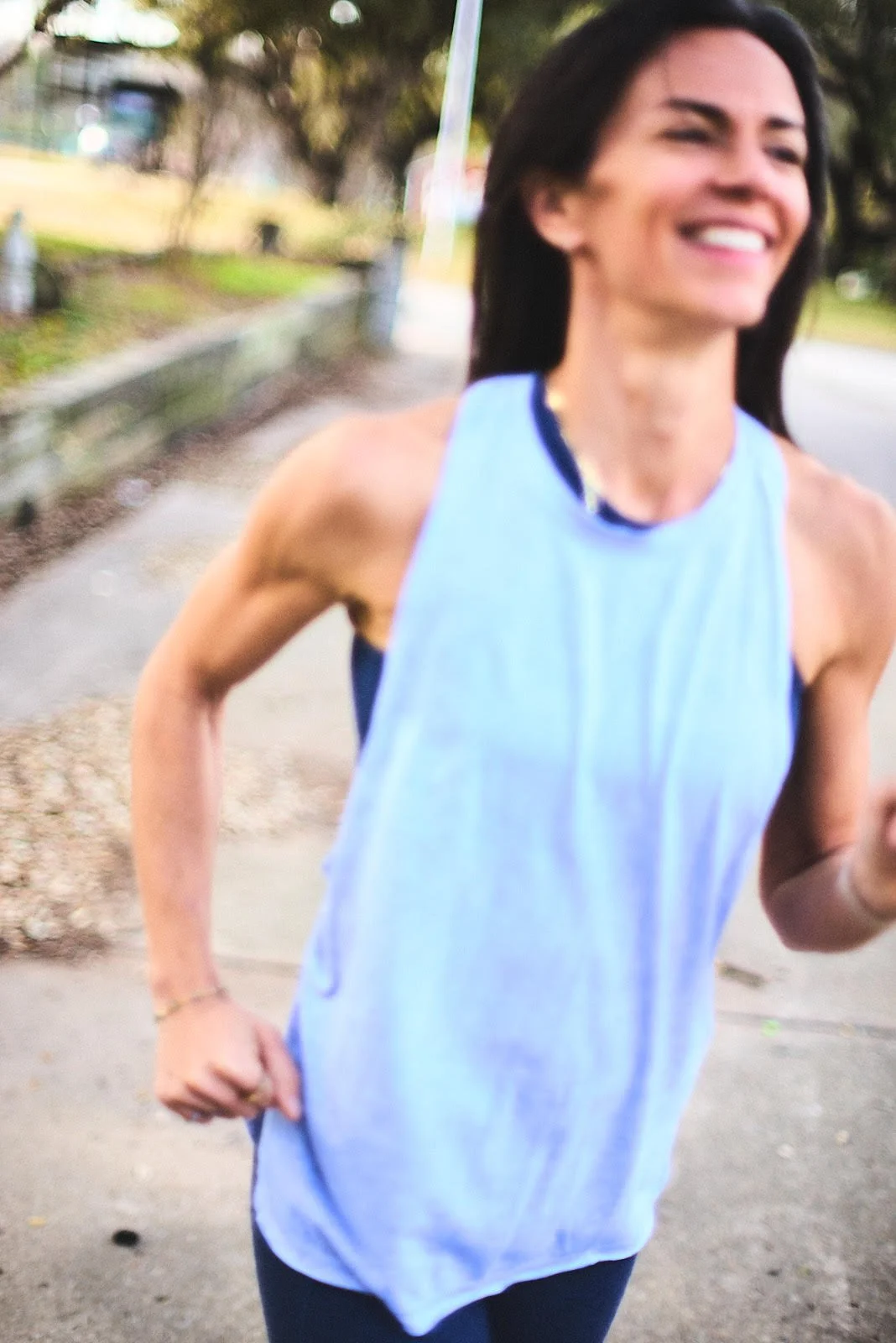 A woman jogging outdoors on a sidewalk, smiling, wearing a light blue athletic tank top and dark pants.