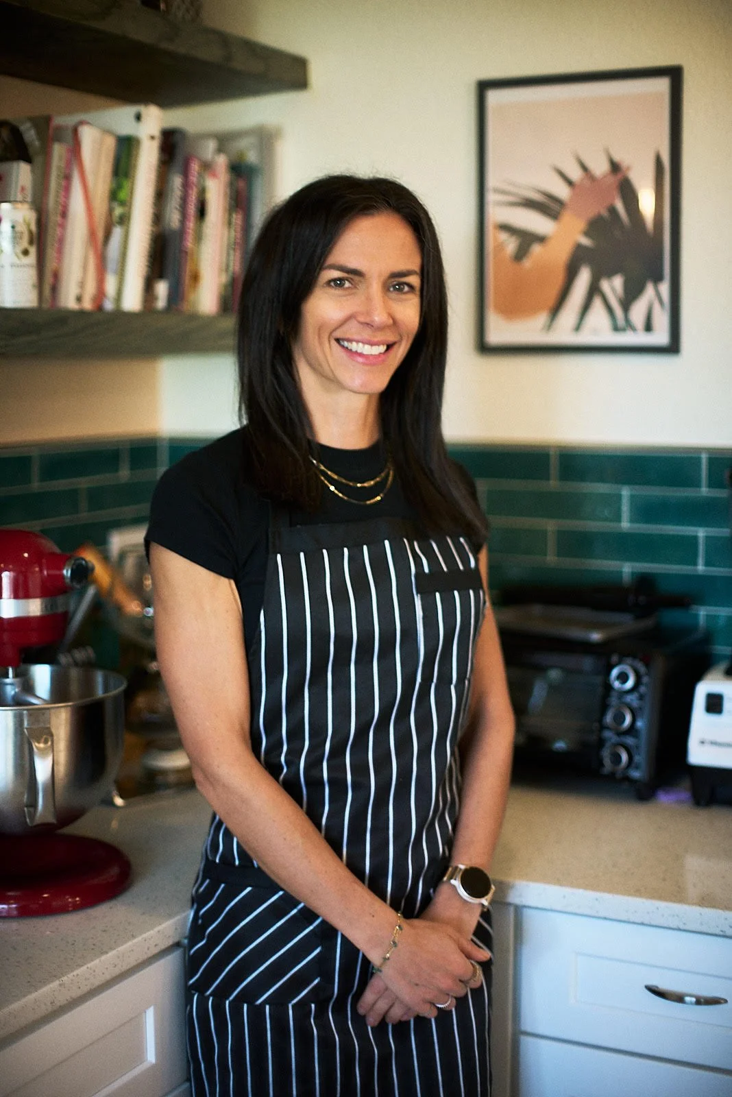 A woman with dark hair smiling, wearing a black shirt and a black and white striped apron, standing in a kitchen with green tile backsplash, books on a shelf, and various kitchen appliances.