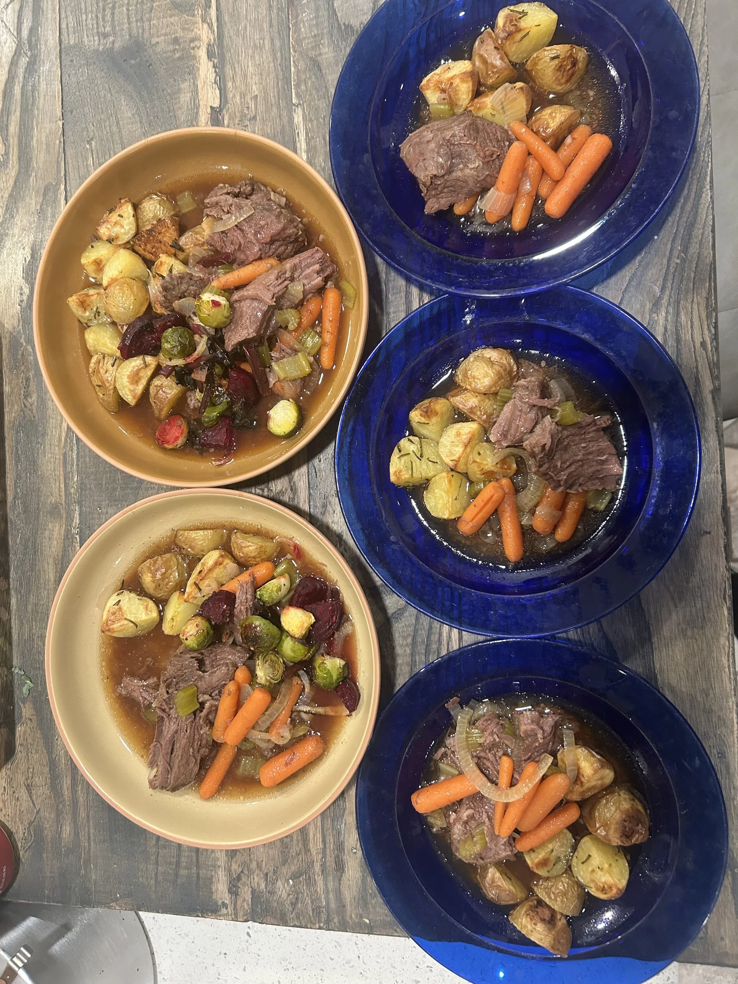 Four plates of cooked meat with carrots, potatoes, onions, and beets, on a wooden table.