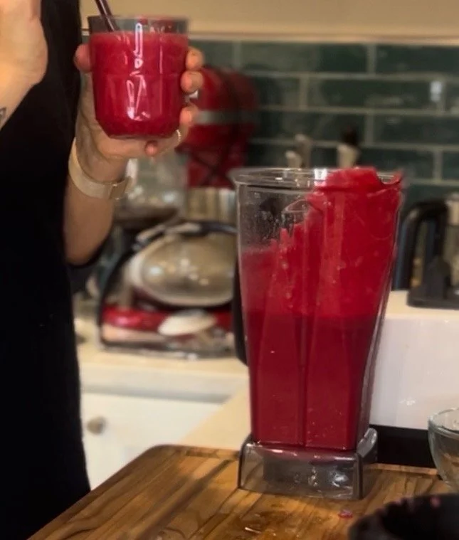 Person holding a glass of red smoothie next to a large blender filled with more red smoothie in a kitchen.