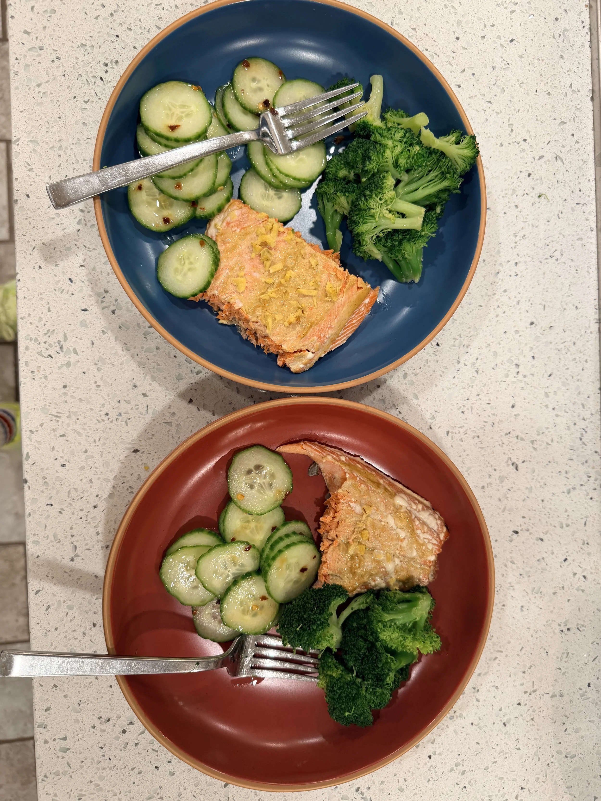 Two colorful bowls on a white speckled countertop, each containing sliced cucumbers, a piece of baked salmon with garlic and lemon, and steamed broccoli.
