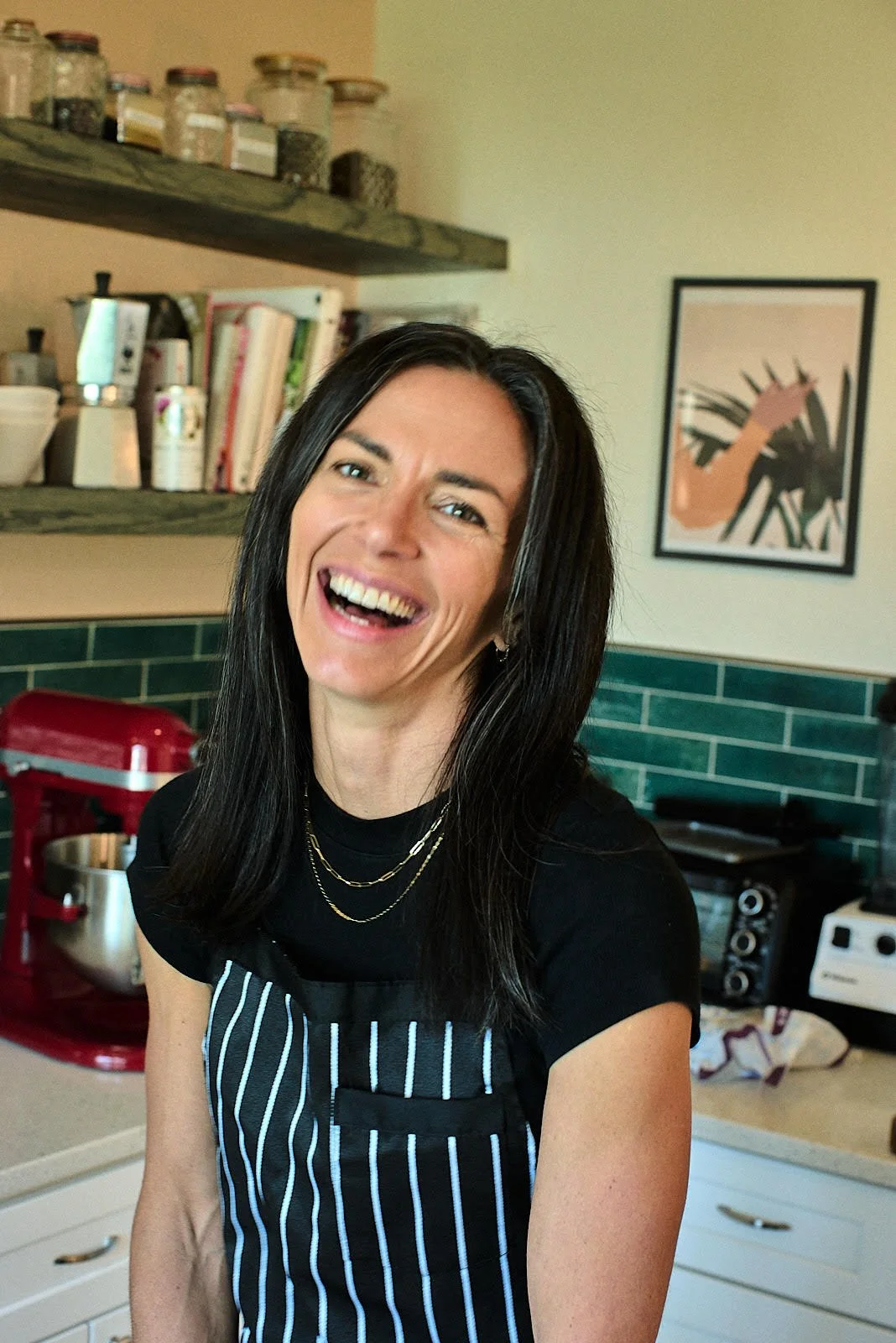 Woman in a black striped apron smiling in a kitchen with green tiles, shelves with jars, and a framed artwork on the wall.