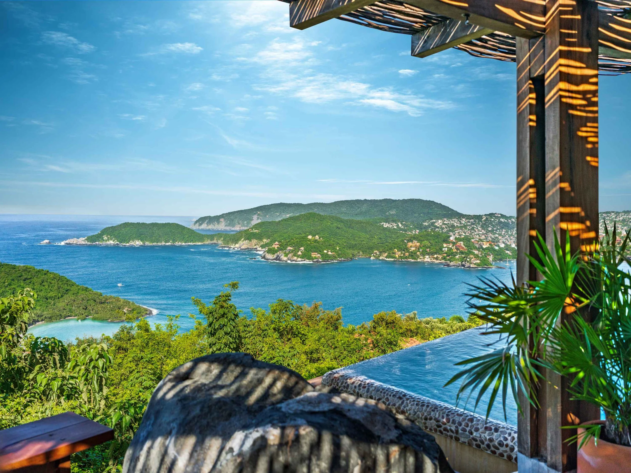 Beautiful view of the bay  of Zihuatanejo with lush green islands and a seaside town on the horizon, seen from a shaded outdoor terrace with tropical plants and decorative wooden and stone accents. las Gatas beach panoramic. playa la ropa.