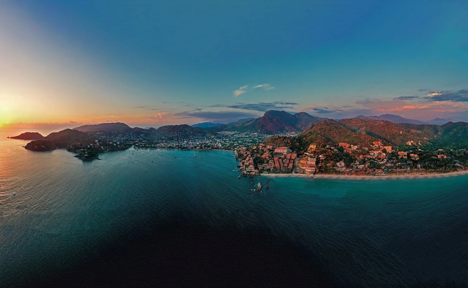 Aerial view of a coastal town at sunset, with mountains in the background, boats in the water, and a beach in the foreground.
