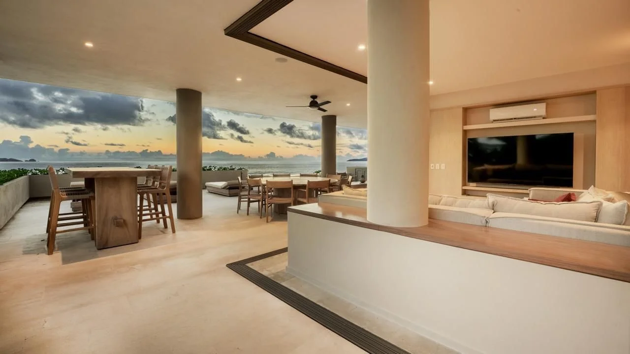 Modern open-concept living space with an outdoor balcony overlooking the ocean at sunset. Features include a dining table, seating area, ceiling fan, and a large TV with a beige sectional sofa.