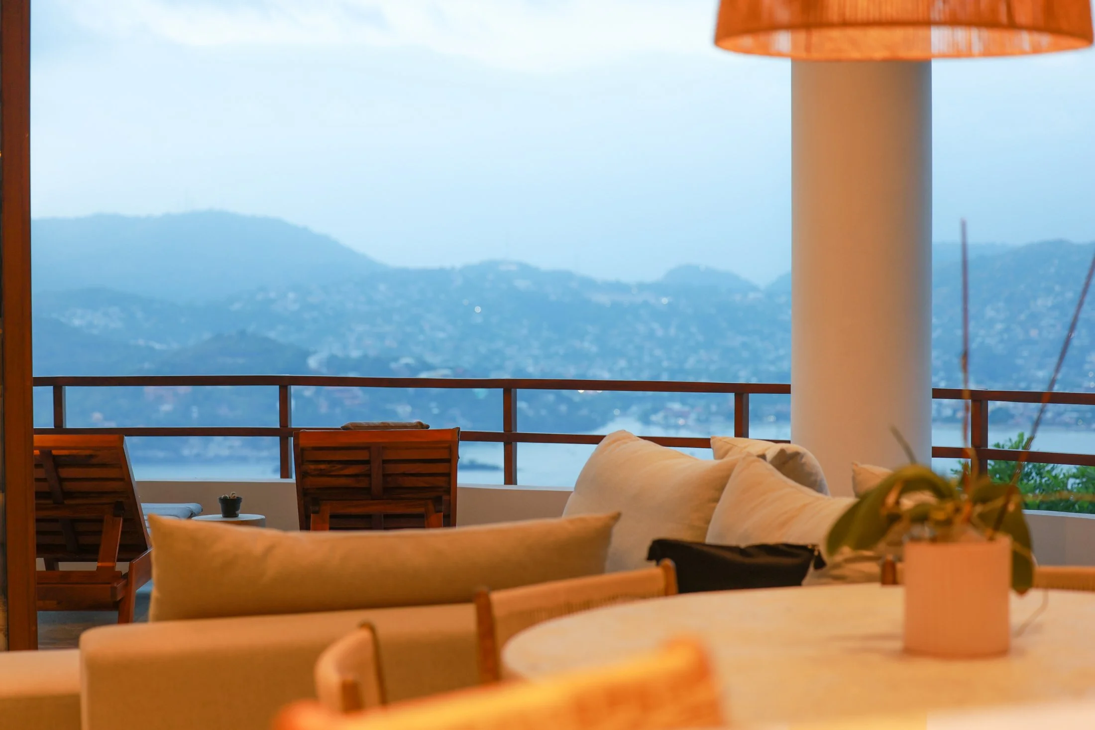 Indoor seating area with a view of mountains and water, featuring beige sofas, a round table with a potted plant, and wooden chairs along a balcony railing.