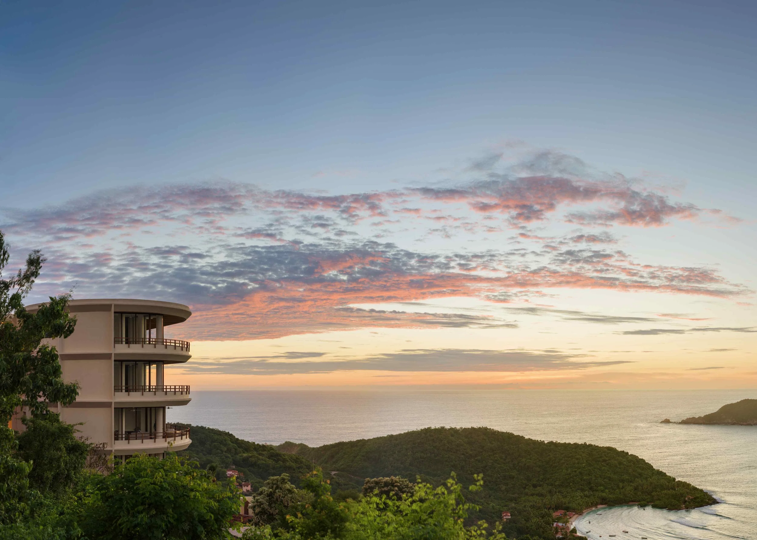 A building with curved balconies overlooking green hills, the ocean, and a colorful sunset sky with clouds.