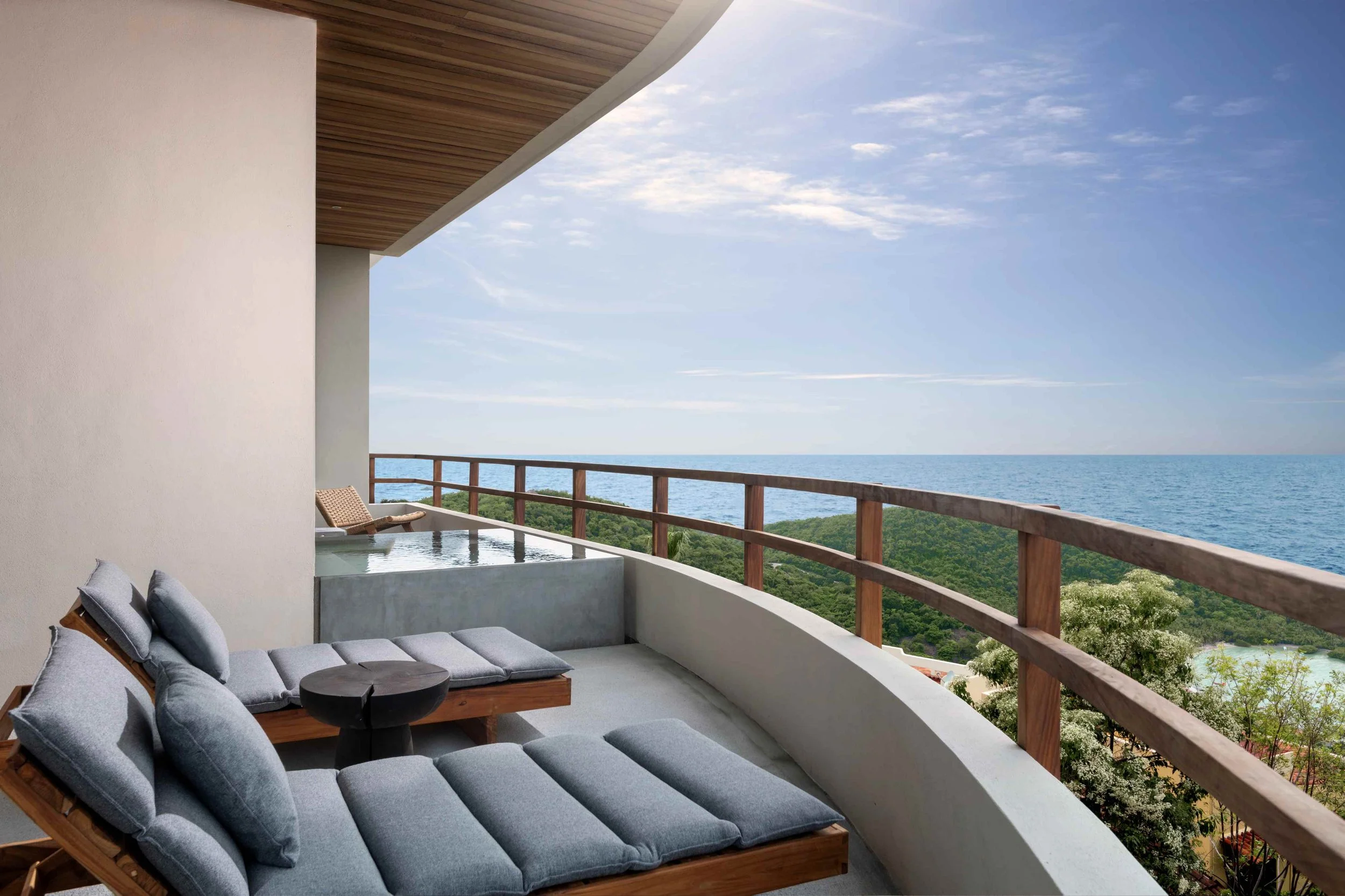 Balcony with gray cushioned lounge chairs, a small black table, wooden railing, overlooking green trees, ocean, and a blue sky with clouds.