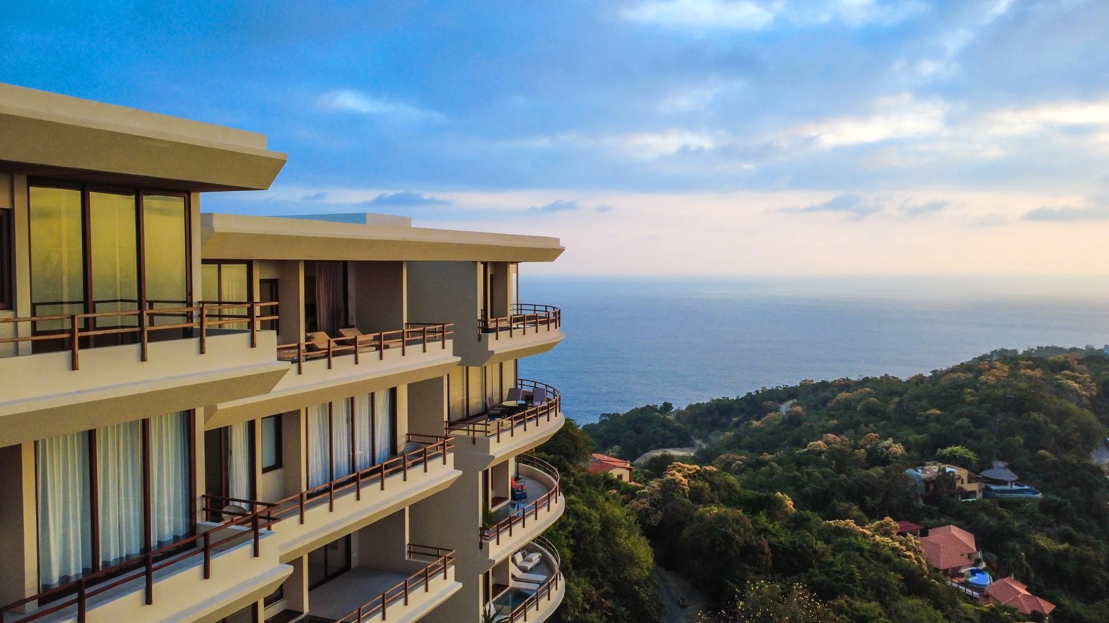 A multi-story building with balconies overlooking lush green trees and the ocean under a partly cloudy sky.