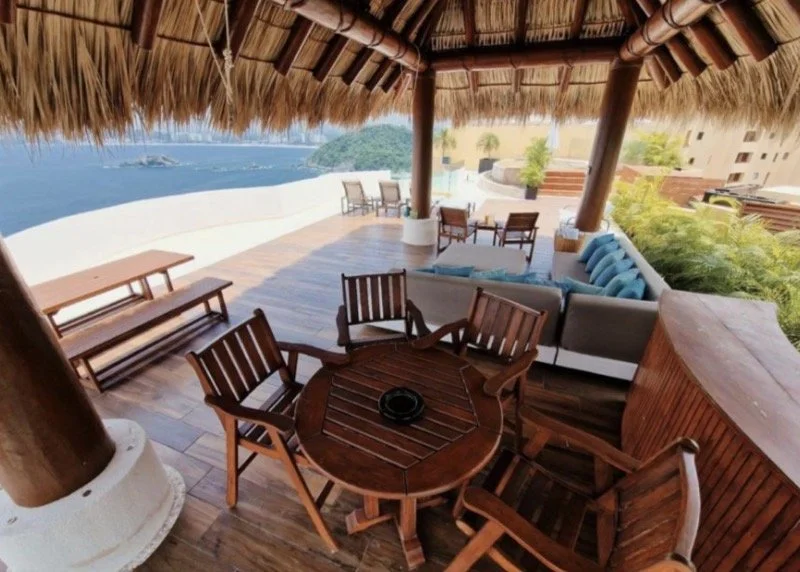 A rooftop patio of a penthouse in ixtapa zihuatanejo with a thatched roof, outdoor furniture including a round wooden table with chairs, a cushioned sofa, and additional chairs, overlooking a scenic view of the pacific ocean and distant hills.