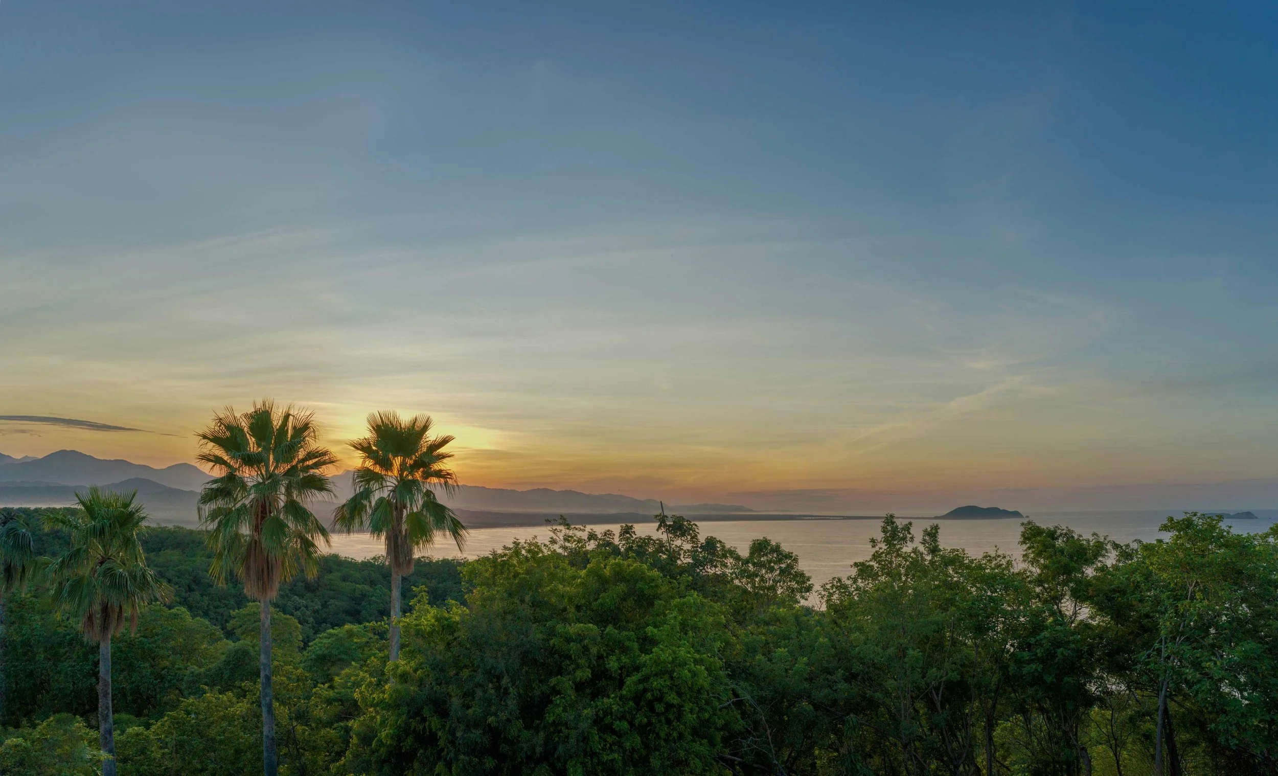 Sunset over a body of water with mountains in the background, palm trees and lush green trees in the foreground.