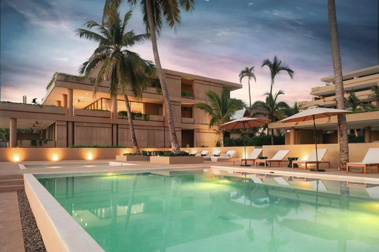 A luxurious hotel pool area at sunset with palm trees, lounge chairs, large umbrellas, and modern buildings in the background. Playa Larga Beach Front Villas. Anera.