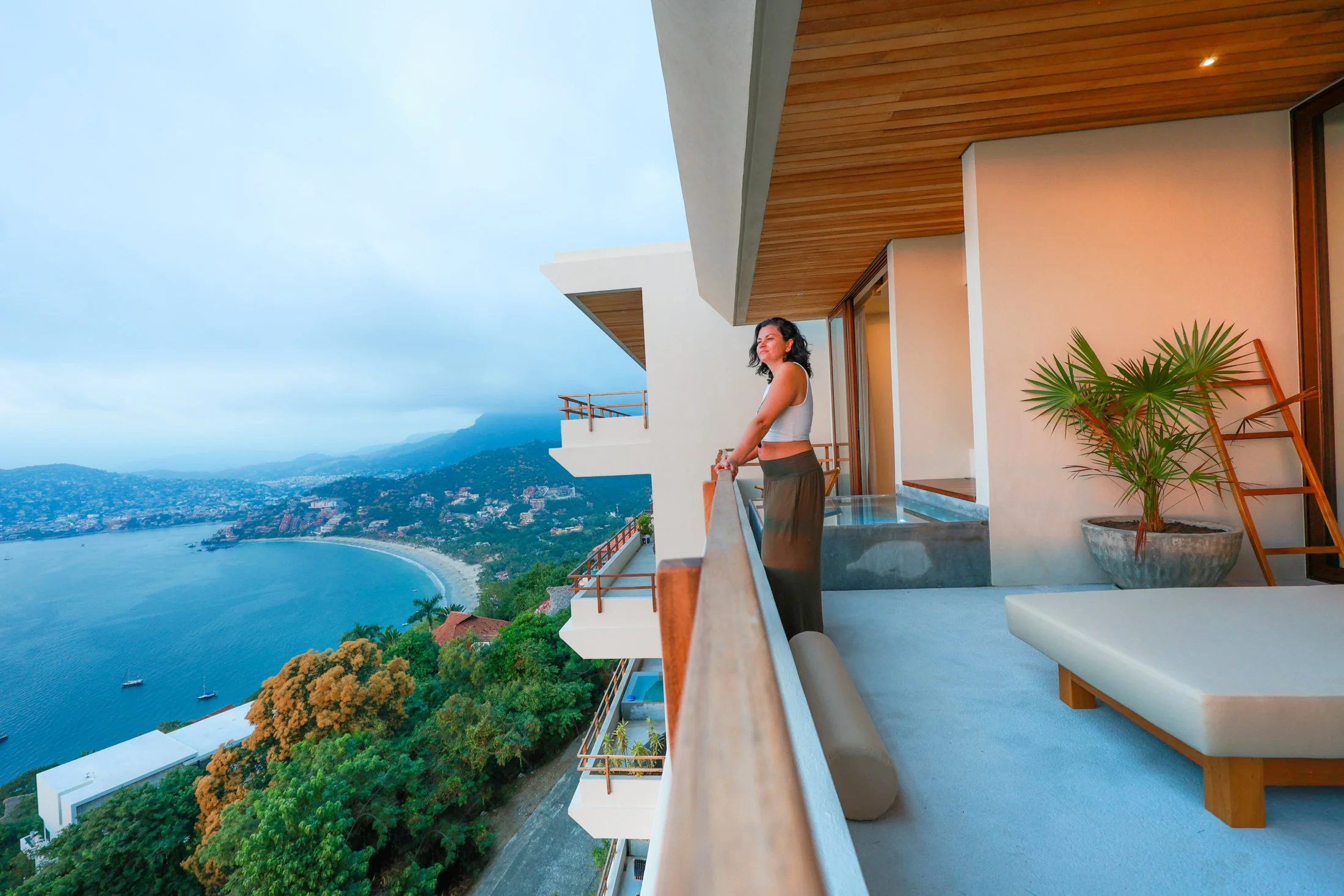 A woman standing on a balcony of a modern building with a scenic view of a bay, mountains, and a cityscape in the distance.