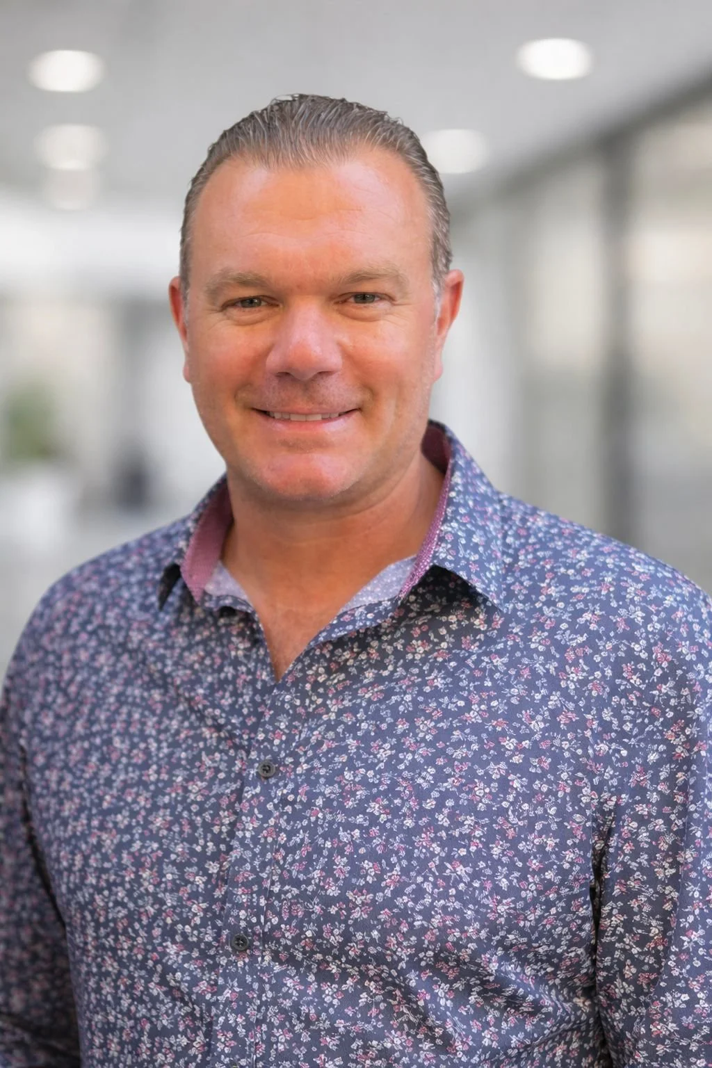 Headshot of a smiling man in a patterned shirt standing in an office corridor.