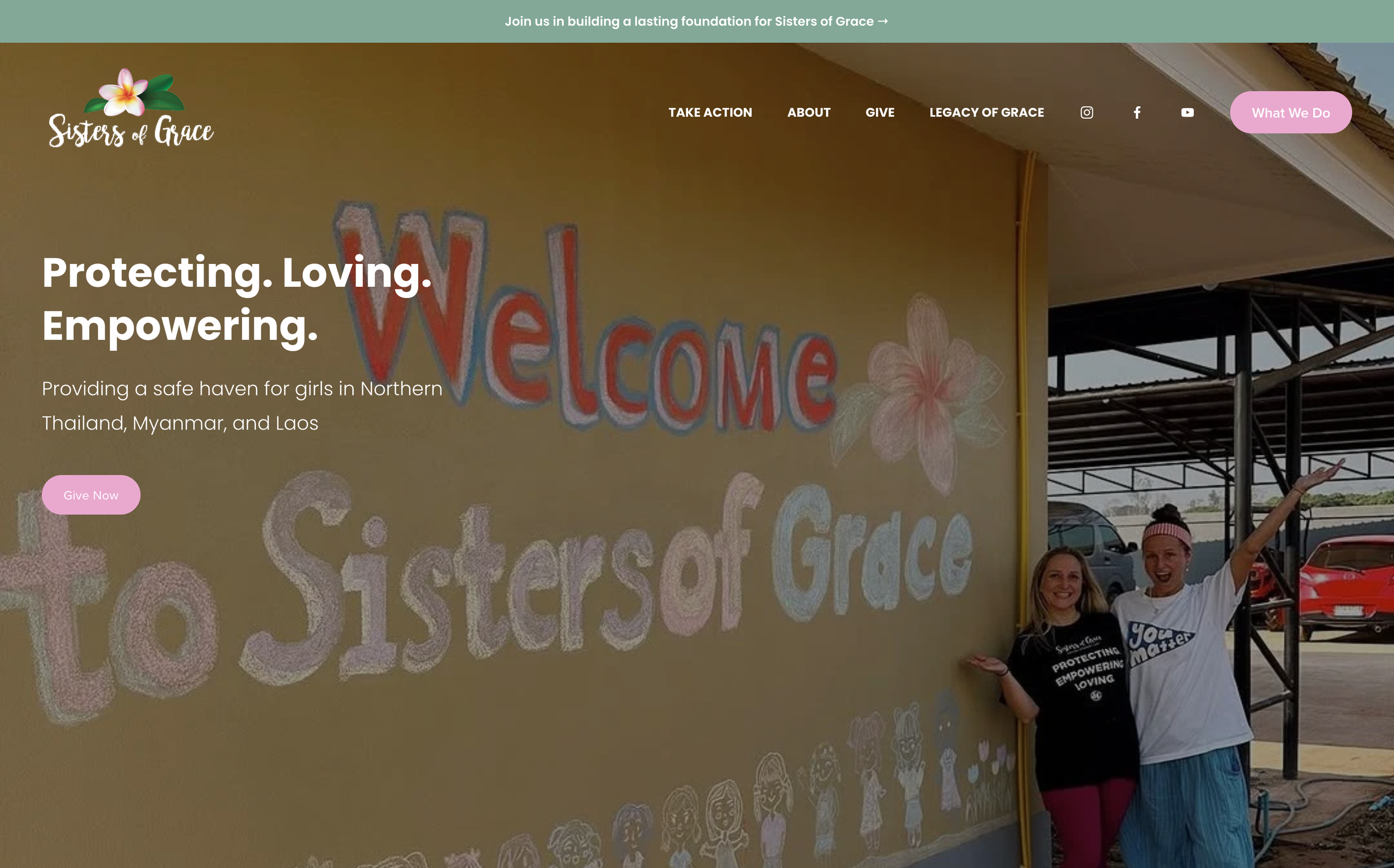 Two women standing in front of a mural on a yellow wall with the words 'Welcome to Sisters of Grace' and a pink flower. One woman is wearing a black T-shirt with white text, and the other is wearing a white T-shirt and a pink headband, both smiling, with one woman raising her arm.