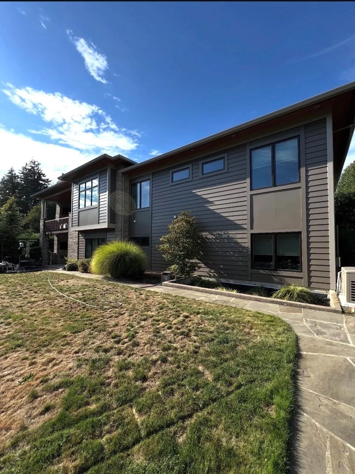 Rear view of a modern two-story house with gray siding, large windows, a stone pillar, and a small patio. The scene has a pathway, lawn, and shrubs under a blue sky with some clouds.