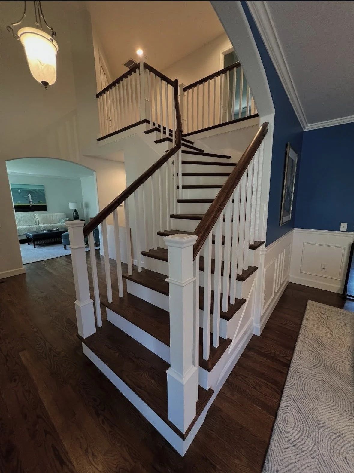 Interior staircase with white railings and dark wooden steps in a home, leading to an upper floor with a blue accent wall and a living room visible in the background.