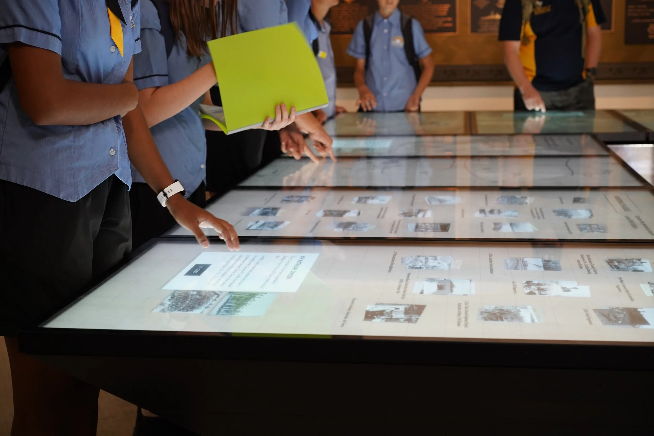 Group of students in blue shirts looking at and interacting with large touchscreen display in a museum or educational setting.