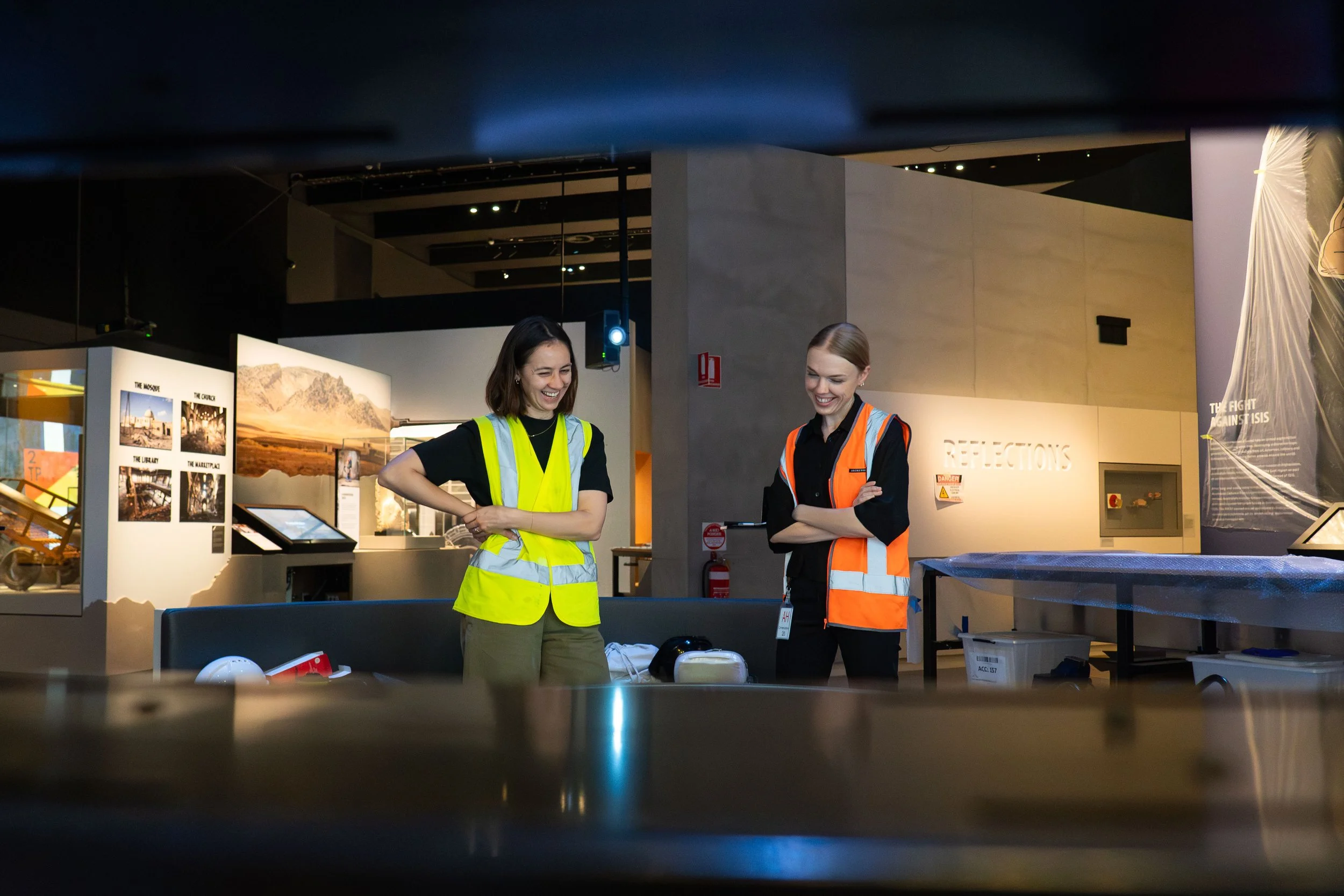 Two women in high-visibility vests standing and talking inside a museum or exhibit space, smiling at each other.