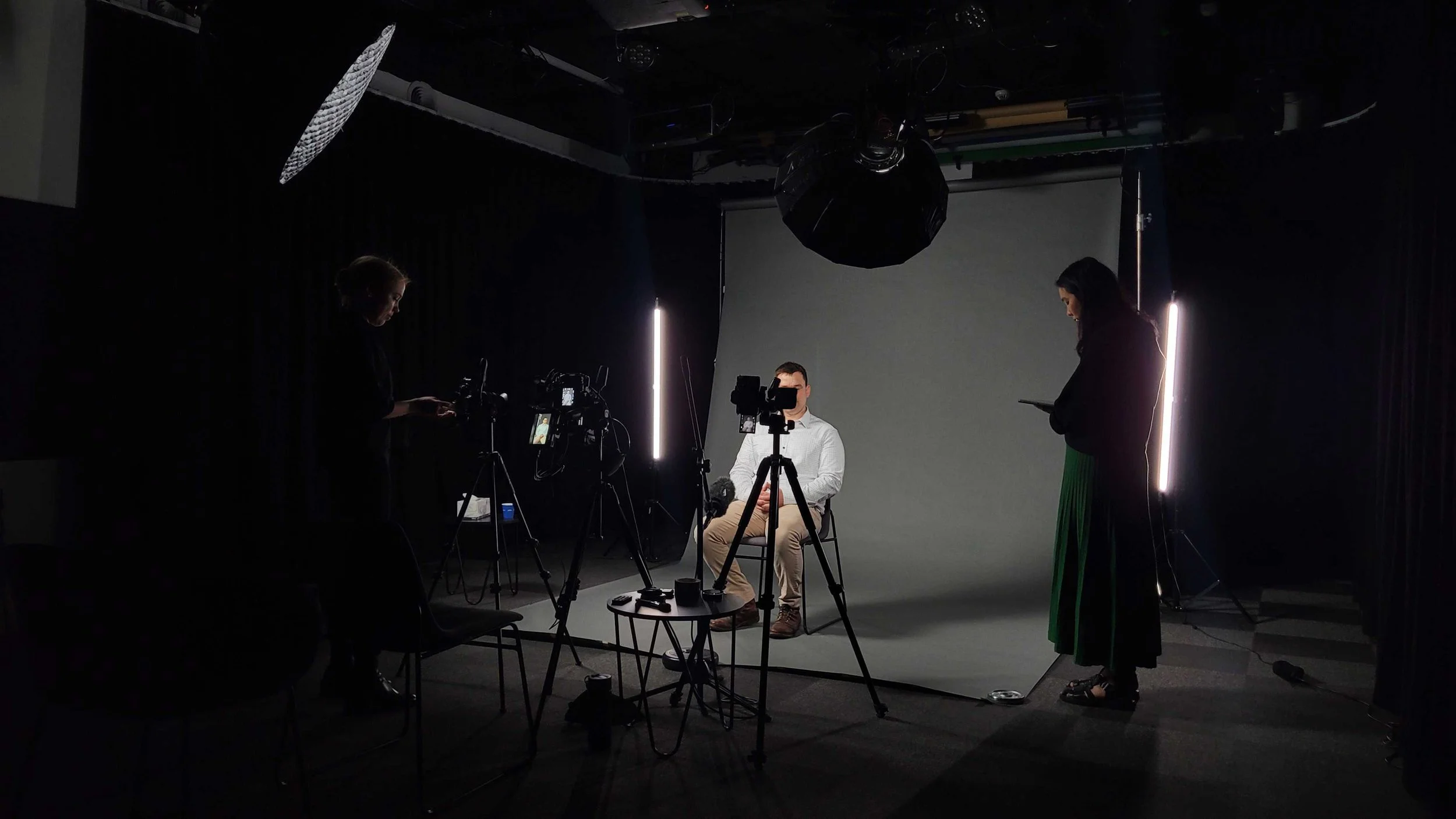 A man is seated in front of a gray backdrop while two women stand nearby in a photography or video studio with equipment, cameras, and lights.