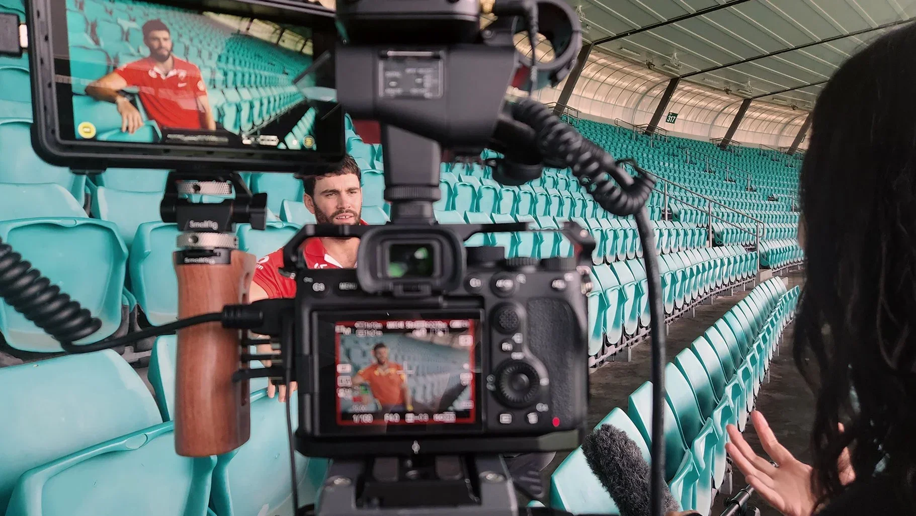 A man in a red sports jersey being recorded by a camera in an empty stadium with teal seats, while a woman with dark hair and dark clothing speaks into a microphone on the right side.