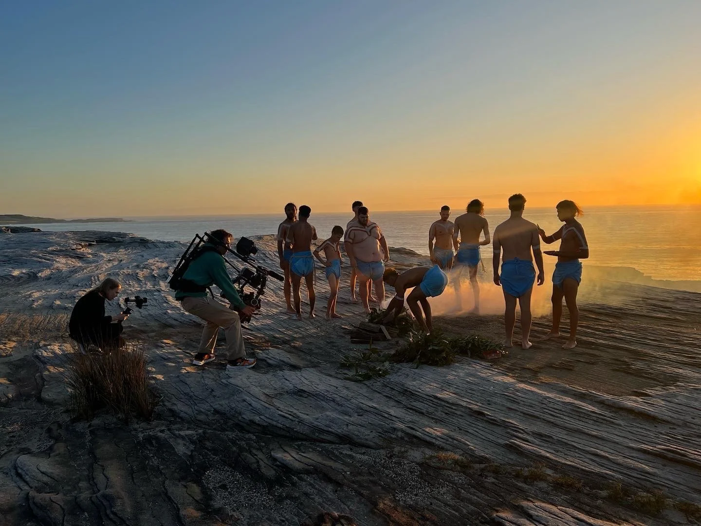 People on a rocky beach filming a scene at sunset, some in blue shorts, with equipment and crew members, capturing a scene near the water.