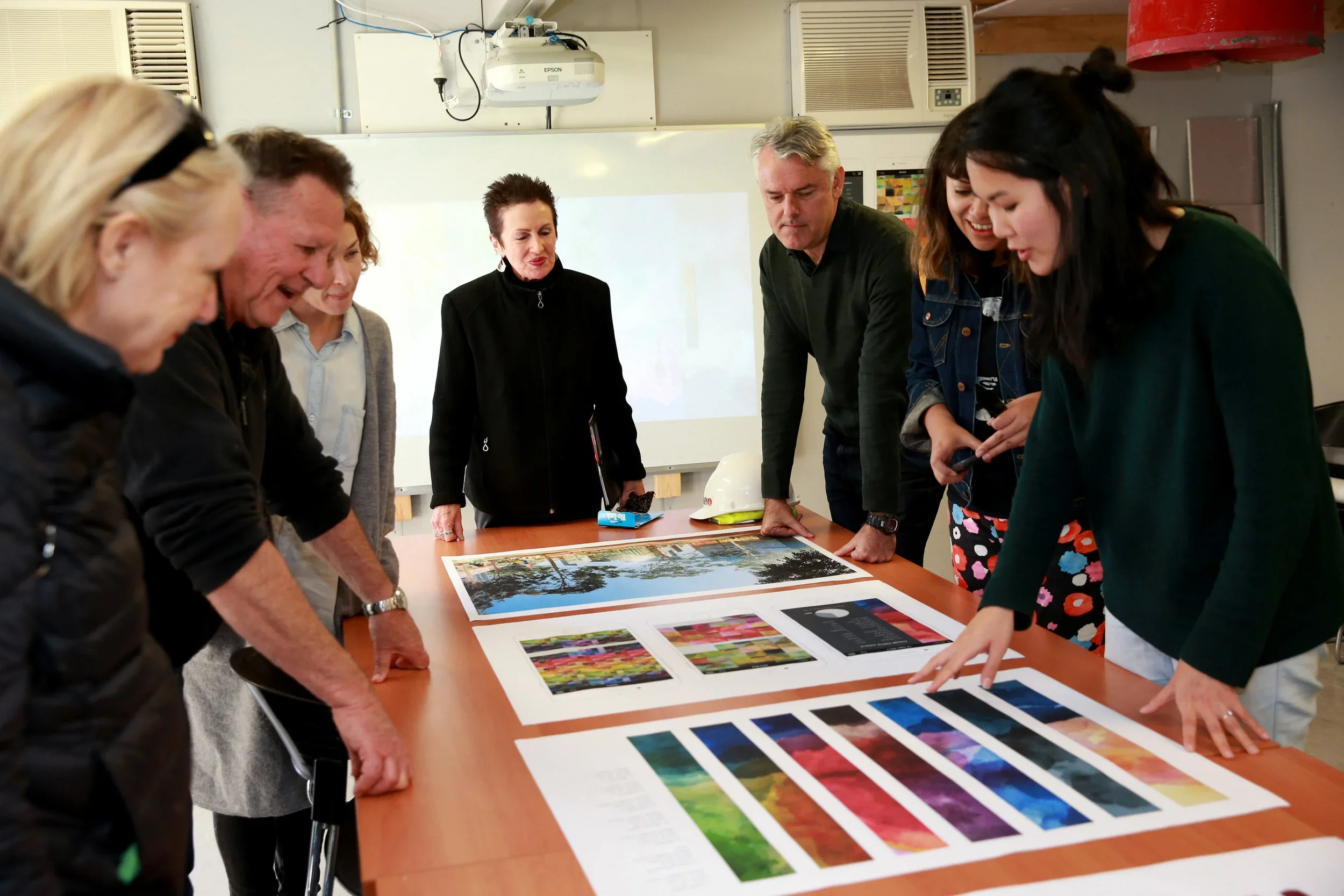 Group of people gathered around a table looking at colorful printed images and art pieces.