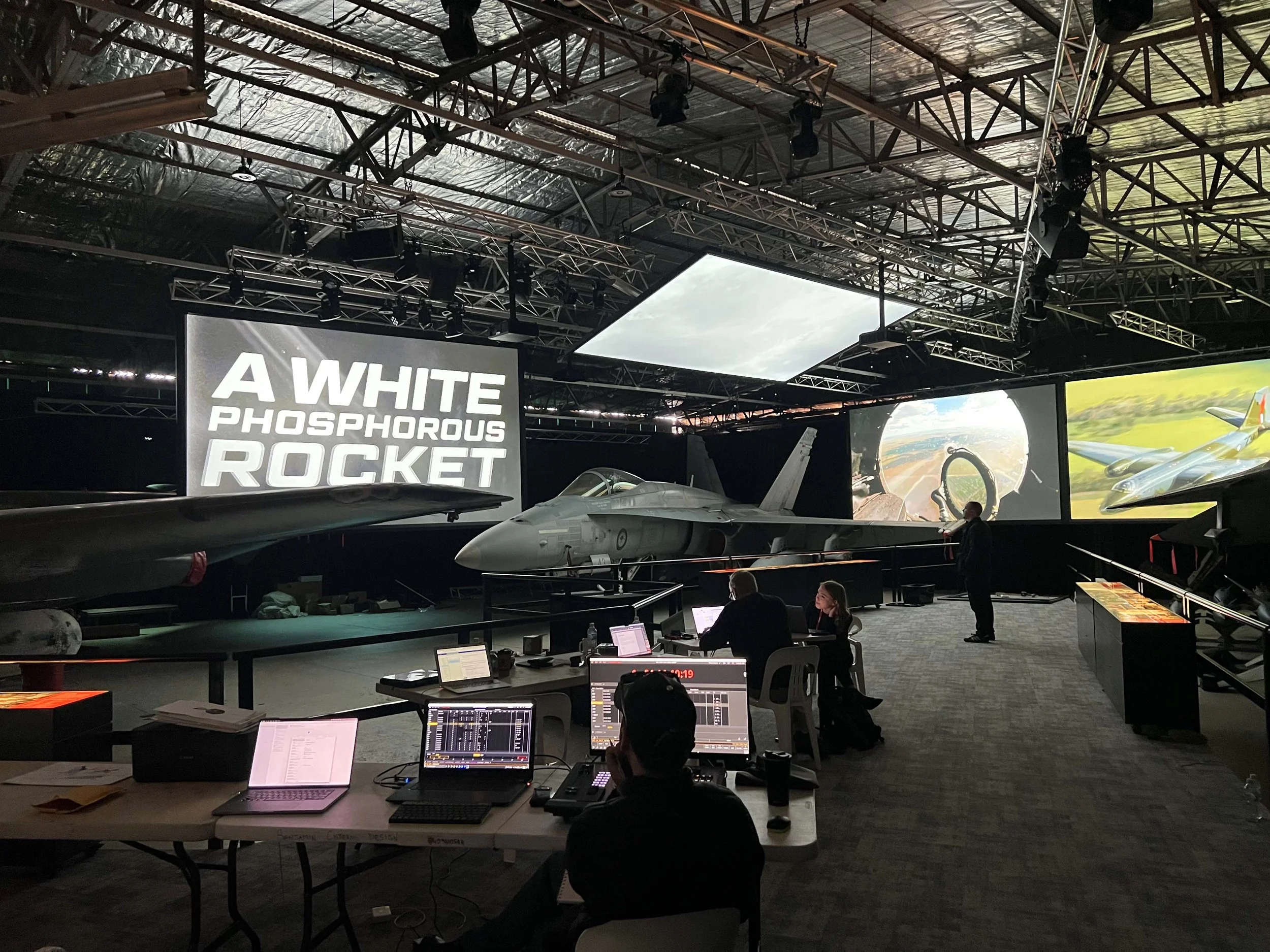 A room with a large display screen showing the text 'A WHITE PHOSPHOROUS ROCKET' and an image of a fighter jet. There is a fighter jet on display in front of the screen, and several people are sitting and working at desks with laptops, while one pers