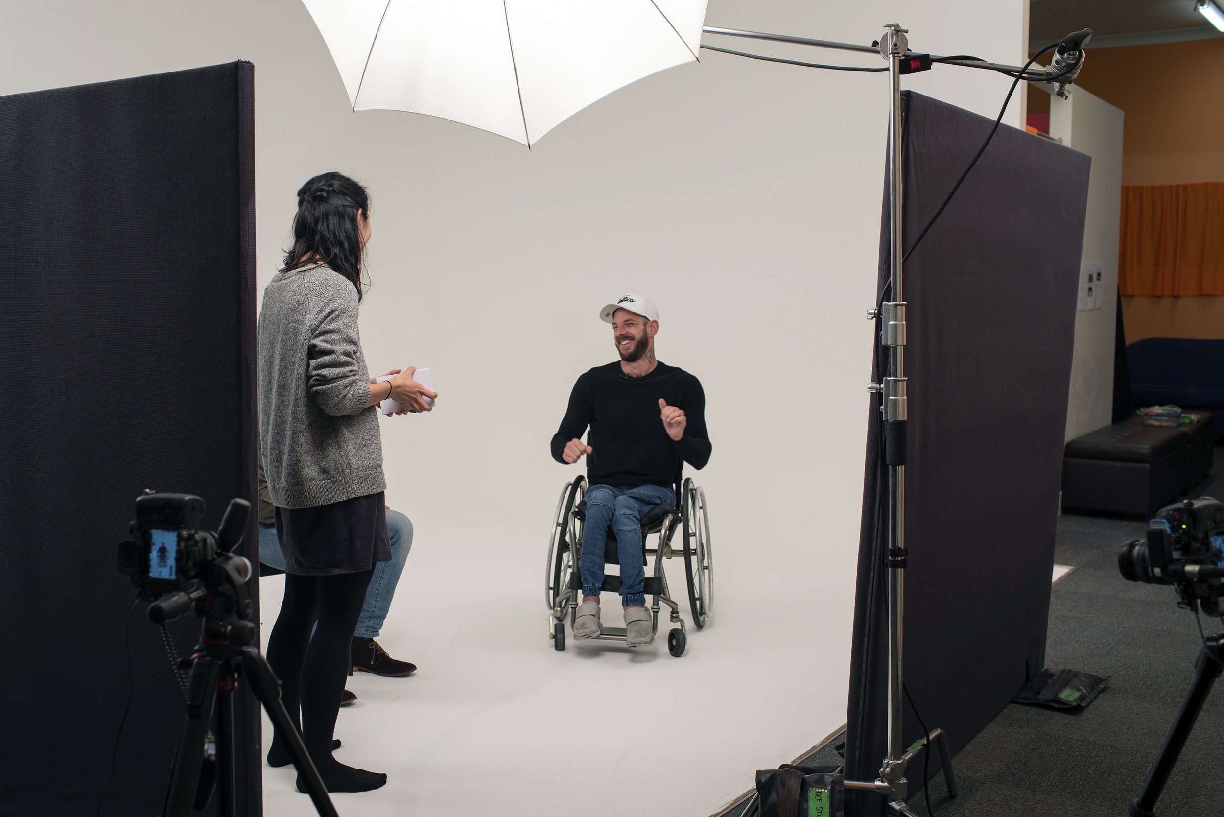 A man in a wheelchair smiling and gesturing during a photo shoot with a woman standing in front of him and holding papers, surrounded by photographic equipment and a white backdrop.