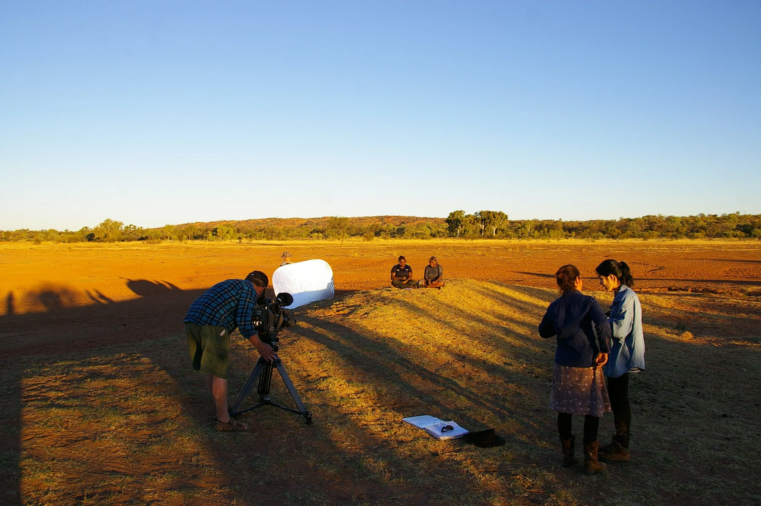 A film crew is filming outdoors on a sunny day in a desert-like area, with two people sitting cross-legged on the ground in the background, and two others standing and talking in the foreground. A person operating a camera is on the left.