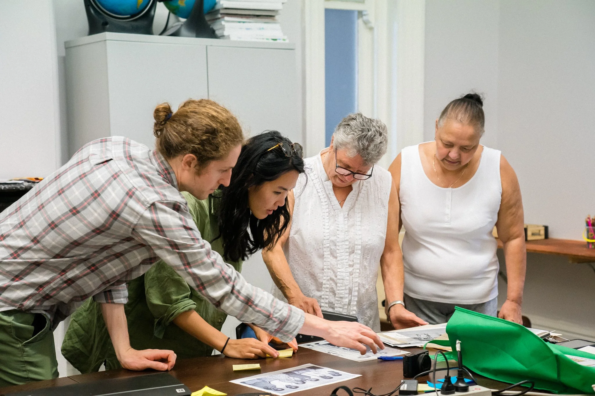 Four women and one man gathered around a table looking at papers and photographs, in an office or classroom setting.
