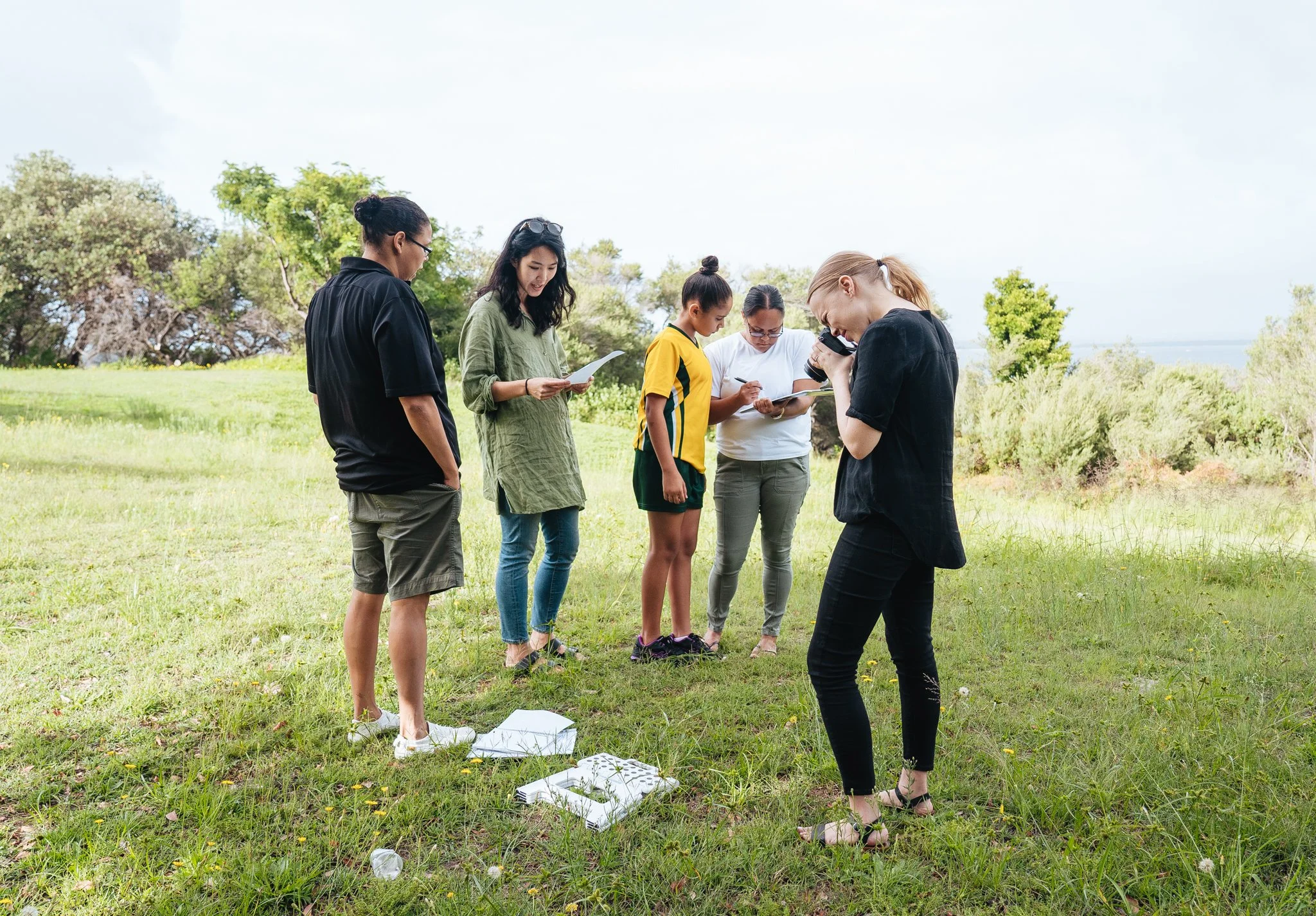 Six women standing on grassy field outdoors, looking at notebooks and camera, with trees and sky in the background.